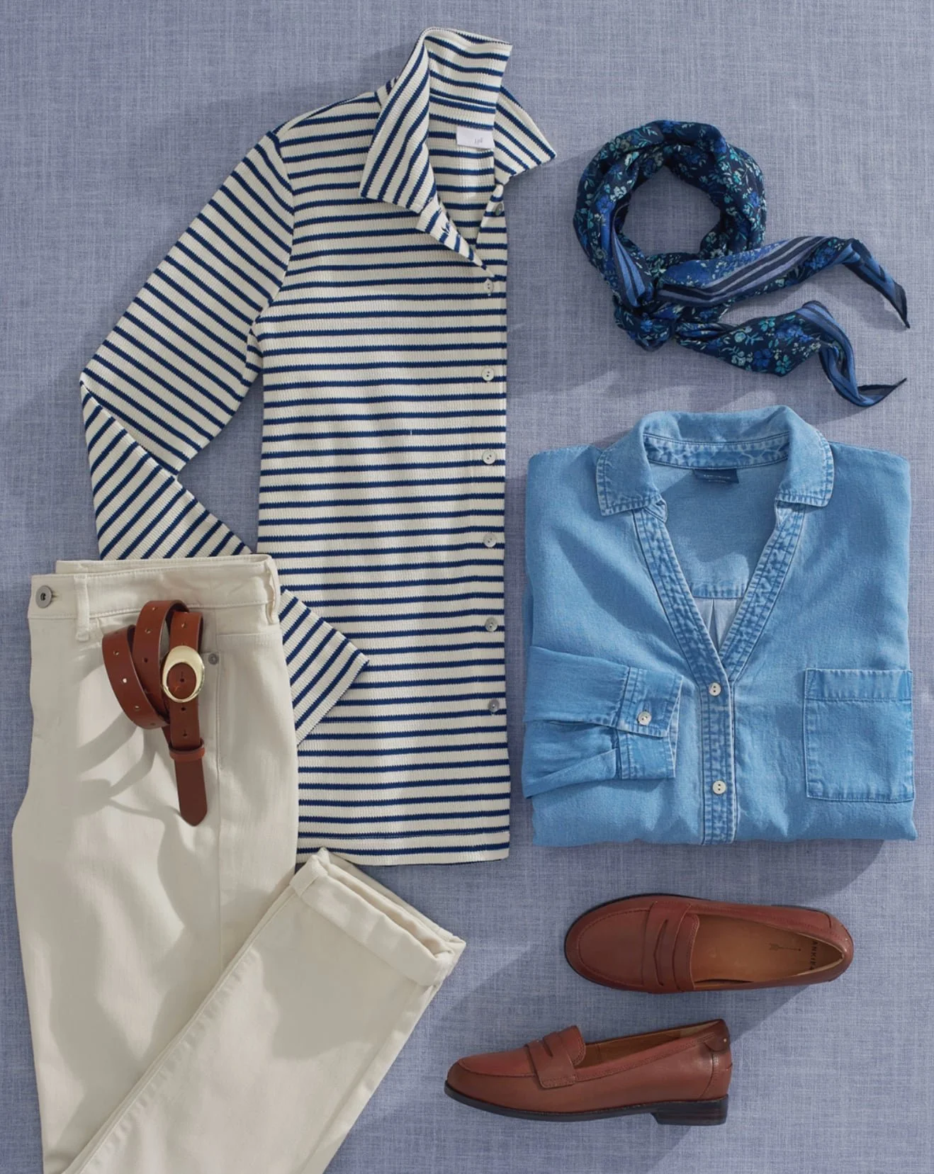 Flat lay of casual women's outfit including a striped long-sleeve shirt, folded light blue denim shirt, white pants with a brown belt, brown loafers, a blue floral scarf, and a pair of brown shoes on a gray fabric background.