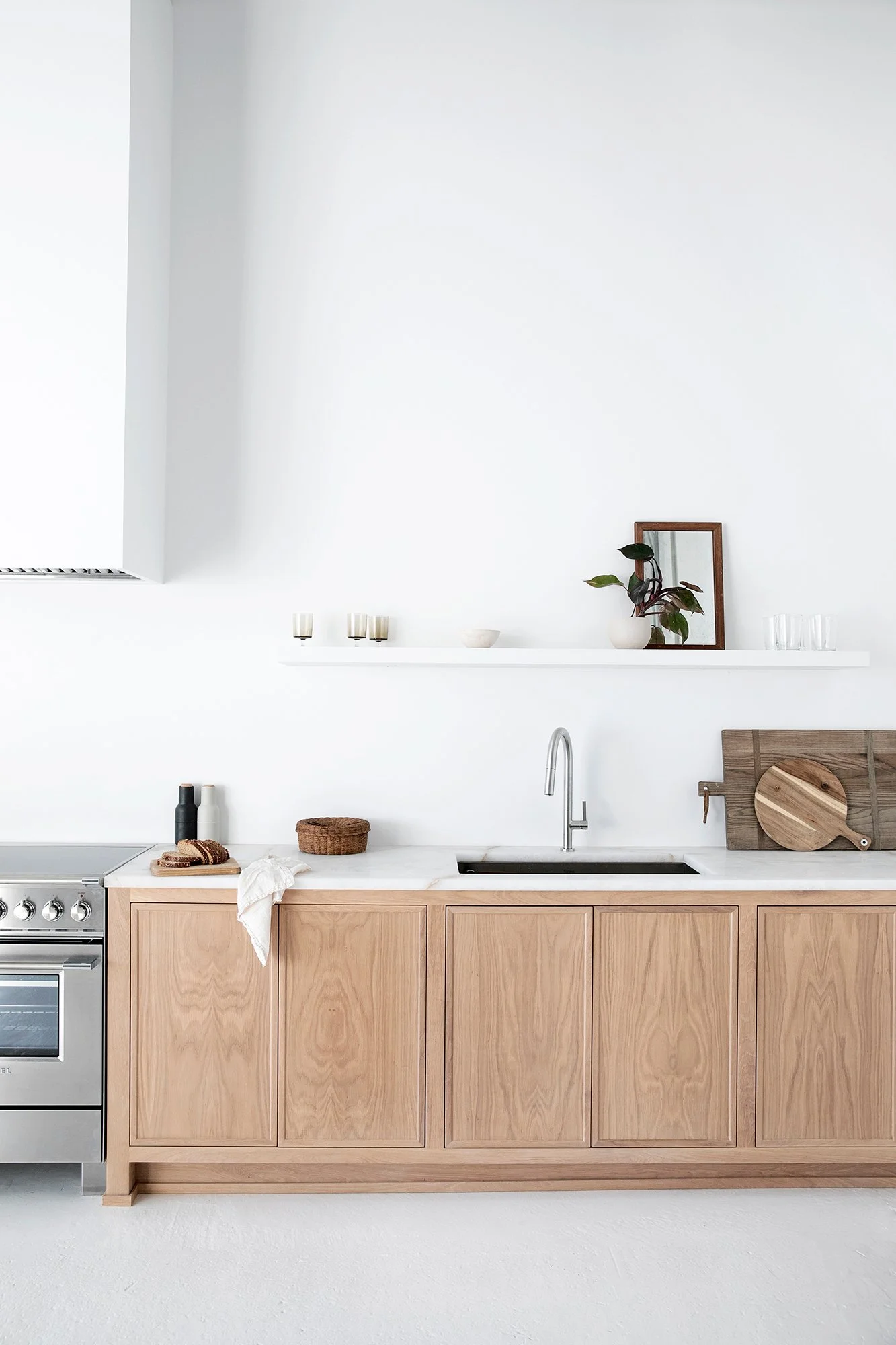 A minimalist kitchen with light wood cabinets, a quartz countertop, a built-in sink, a stovetop, and decor including potted plants, a mirror, candles, and kitchen utensils.
