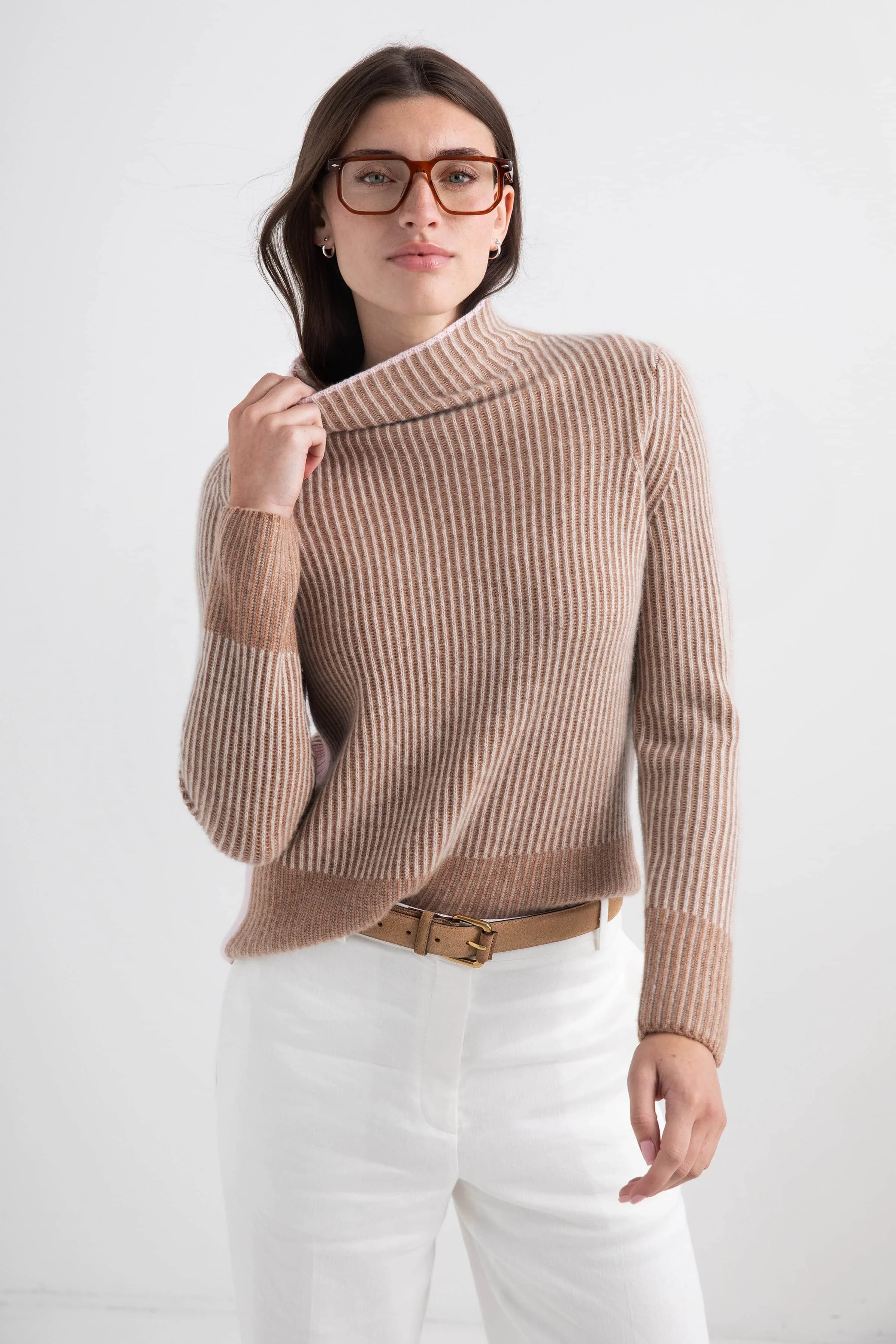 A woman with shoulder-length brown hair, wearing glasses with brown frames, a beige and light pink striped turtleneck sweater, and white pants with a beige belt, standing against a plain white background.
