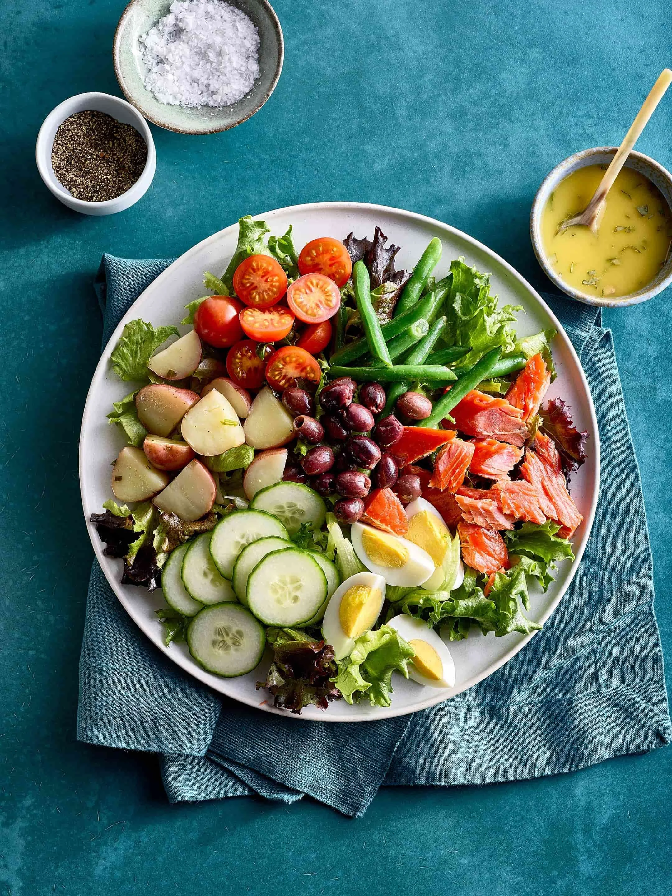 A large white plate with fresh salad ingredients including cherry tomatoes, green beans, cucumber slices, hard-boiled eggs, grapes, and shredded salmon on a teal tablecloth with a folded blue napkin. Around the plate are small bowls of salt, pepper, 
