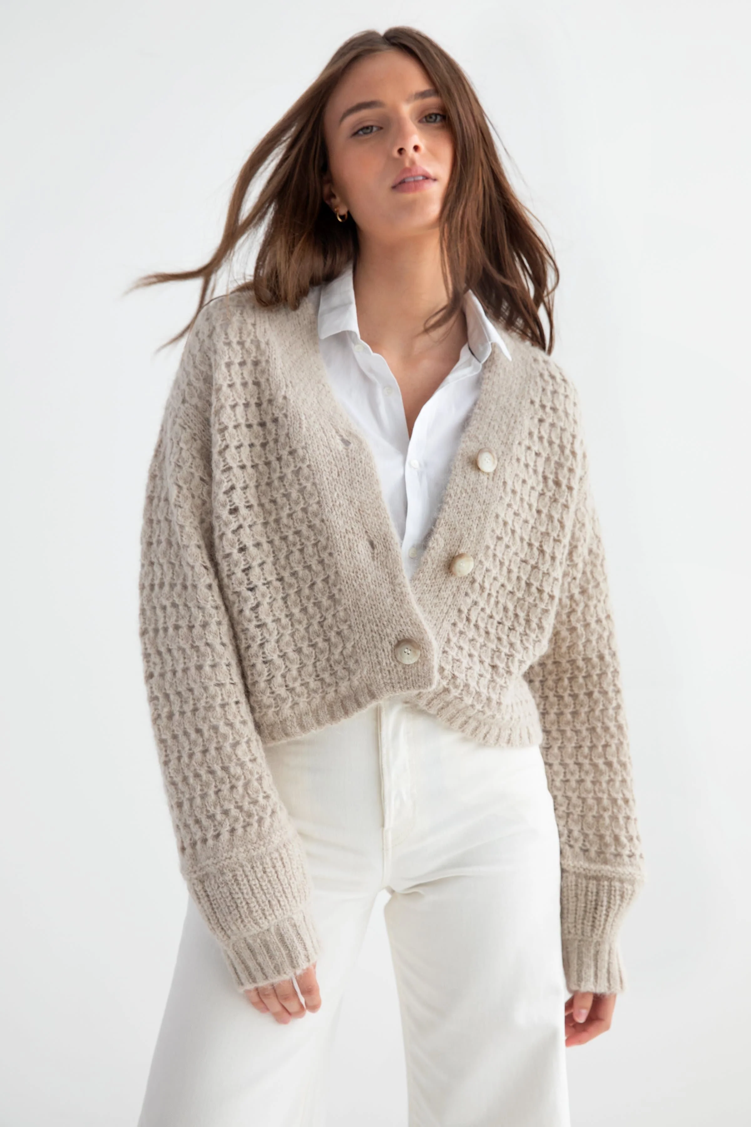 Woman wearing a beige knit cardigan over a white shirt and white pants, with the focus on her upper body and face, standing against a plain white background.
