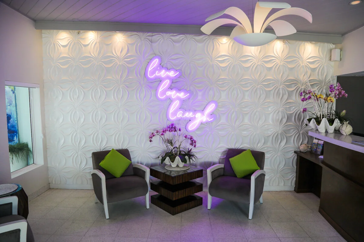 A cozy seating area with two black armchairs with white trim and green pillows, a small dark wood table with a decorative flower arrangement, a white textured wall with a purple neon sign that reads 'live love laugh', and purple orchids in white pots