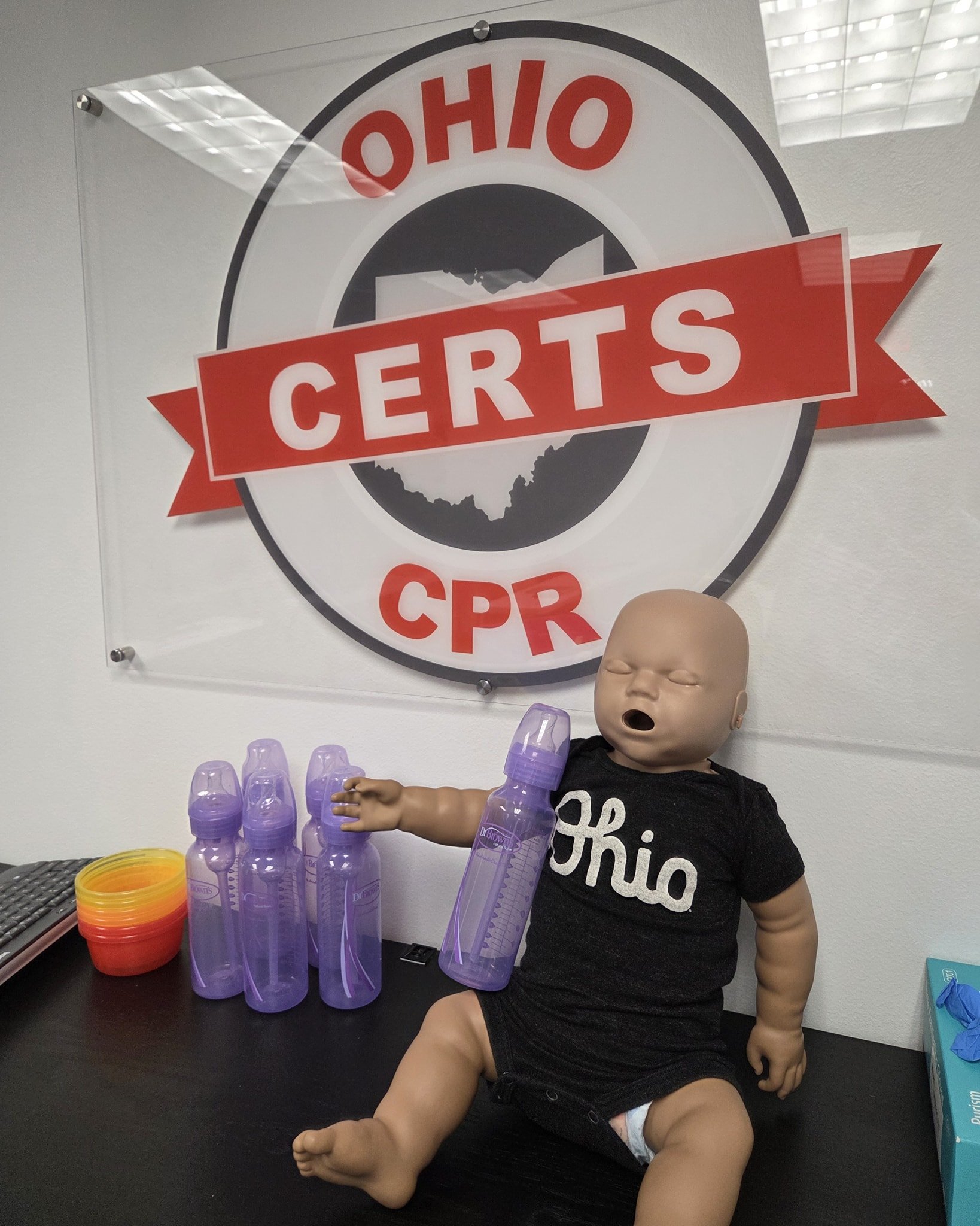 Doll lying on a desk with a sign on the wall behind it that reads "Ohio CERTS CPR". The doll is holding six purple water bottles and is dressed in a black shirt with "Ohio" written on it.