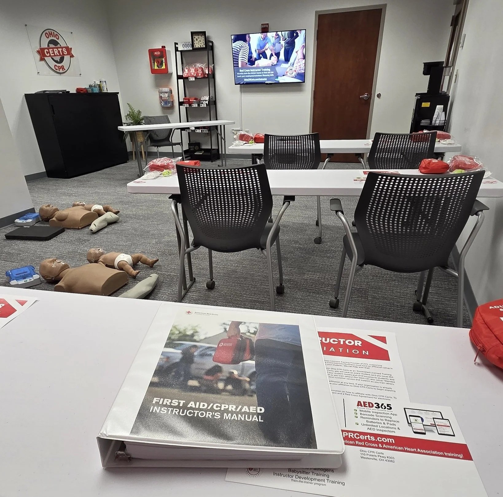 A CPR and first aid training room set up with mannequins on the floor, training materials on the tables, and a monitor displaying instructional content.