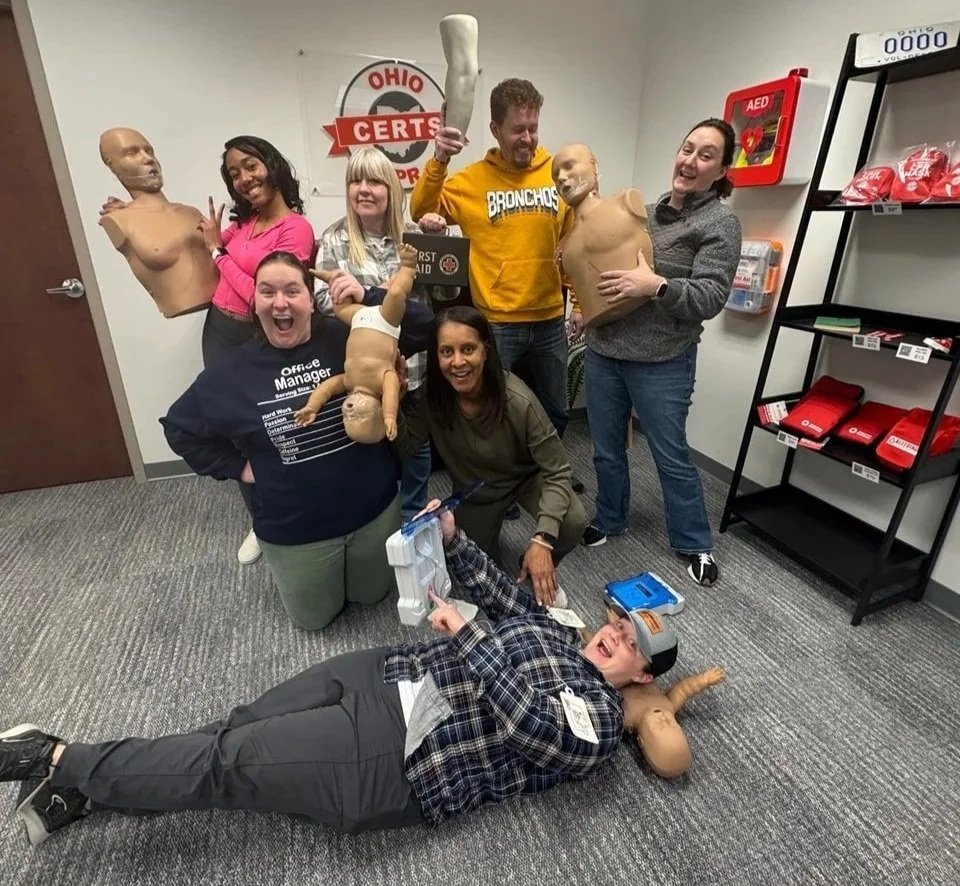 Group of people posing with mannequin kids, CPR training dummies, and first aid awards in a training room with Ohio CERTS sign and emergency supplies.