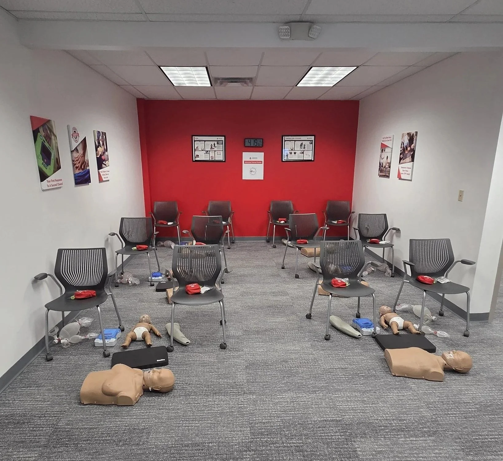 A CPR training room with chairs, mannequins, and medical supplies arranged for practice, with informational posters on the walls and a red wall at the back.
