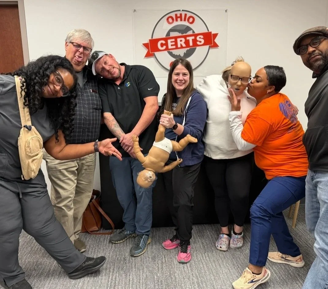 Group of seven diverse adults standing and posing in front of an Ohio CERTs banner, making playful gestures and holding a baby mannequin wearing a diaper.