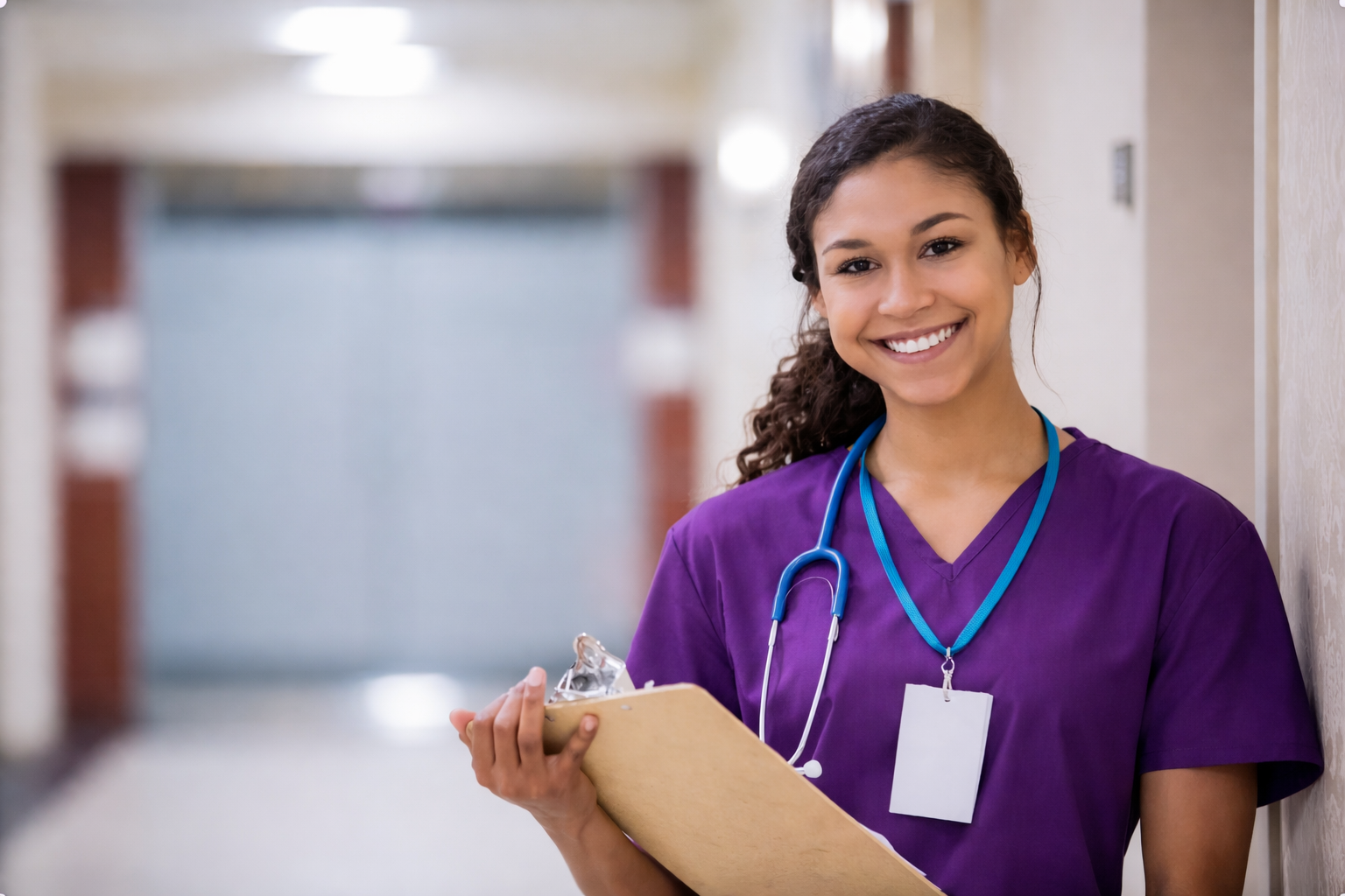 CNA student in purple scrubs standing in a healthcare facility hallway, holding a clipboard and wearing a stethoscope, representing CNA workforce readiness, clinical skills training, and preparation for real-world patient care.