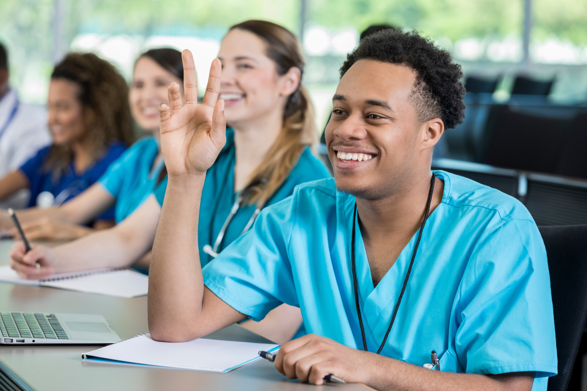 CNA student in teal scrubs raising his hand during classroom training, smiling and engaged alongside fellow healthcare students, representing active learning and clinical skills education in a CNA program.