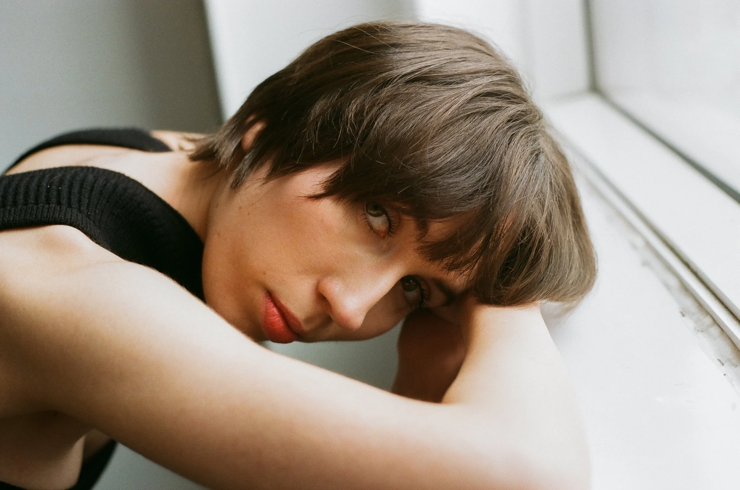 A young woman with short brown hair and light skin rests her head on her arm as she looks at the camera near a window.