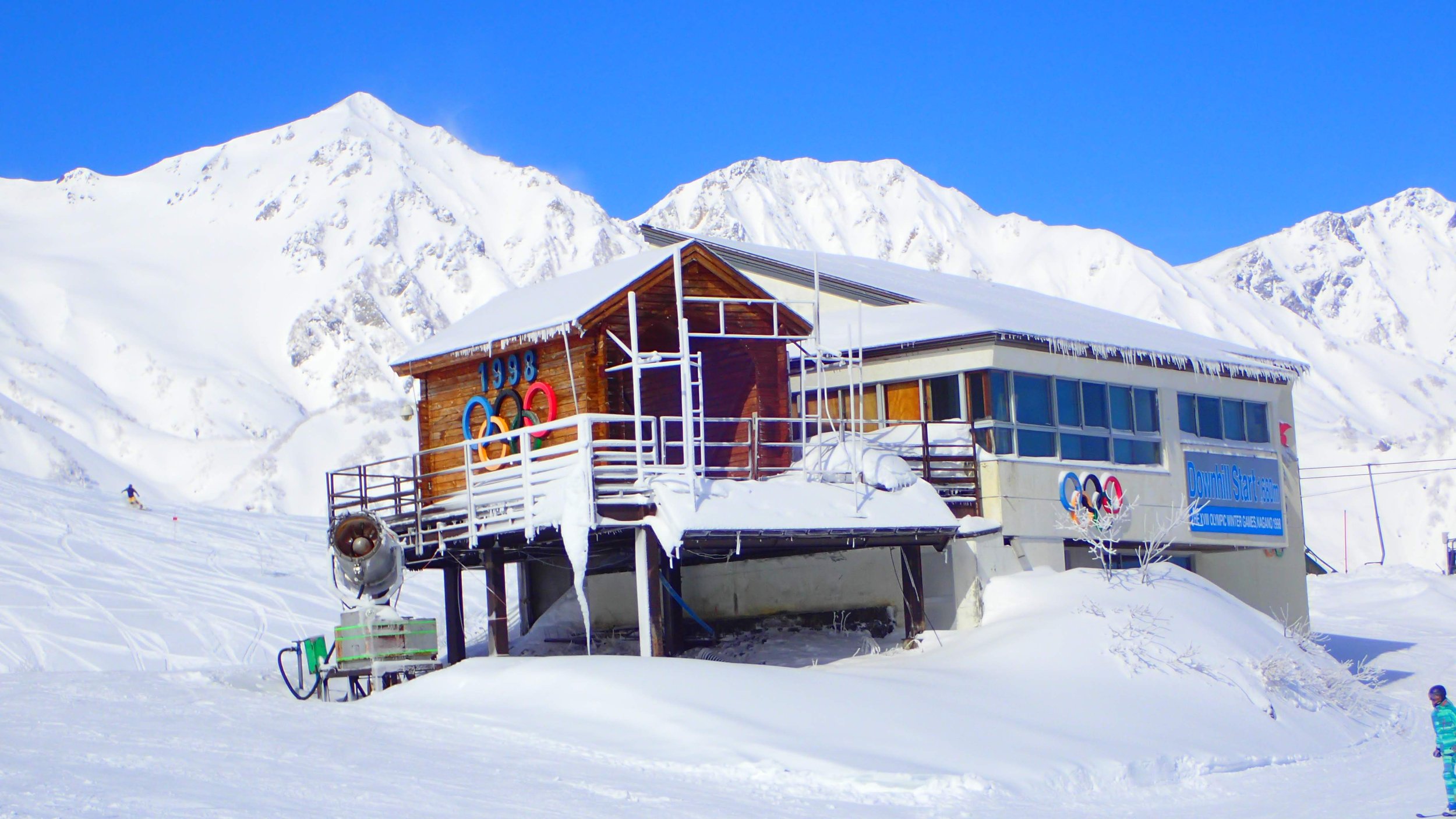 Snow-covered ski resort building in front of tall snowy mountains with clear blue sky, featuring Olympic rings and a sign that reads 'Download Start.' Skiers are visible on the slopes.