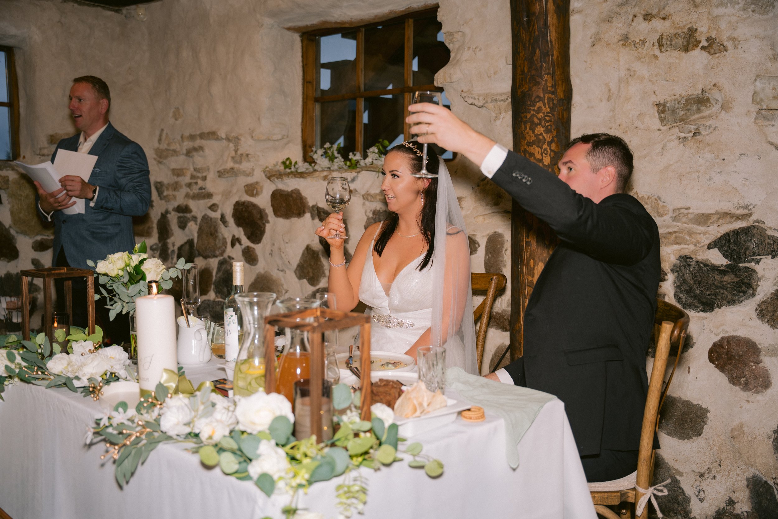A bride and groom at their wedding reception, with the groom raising a toast while the bride holds a wine glass, seated at a decorated table against a rustic stone wall.