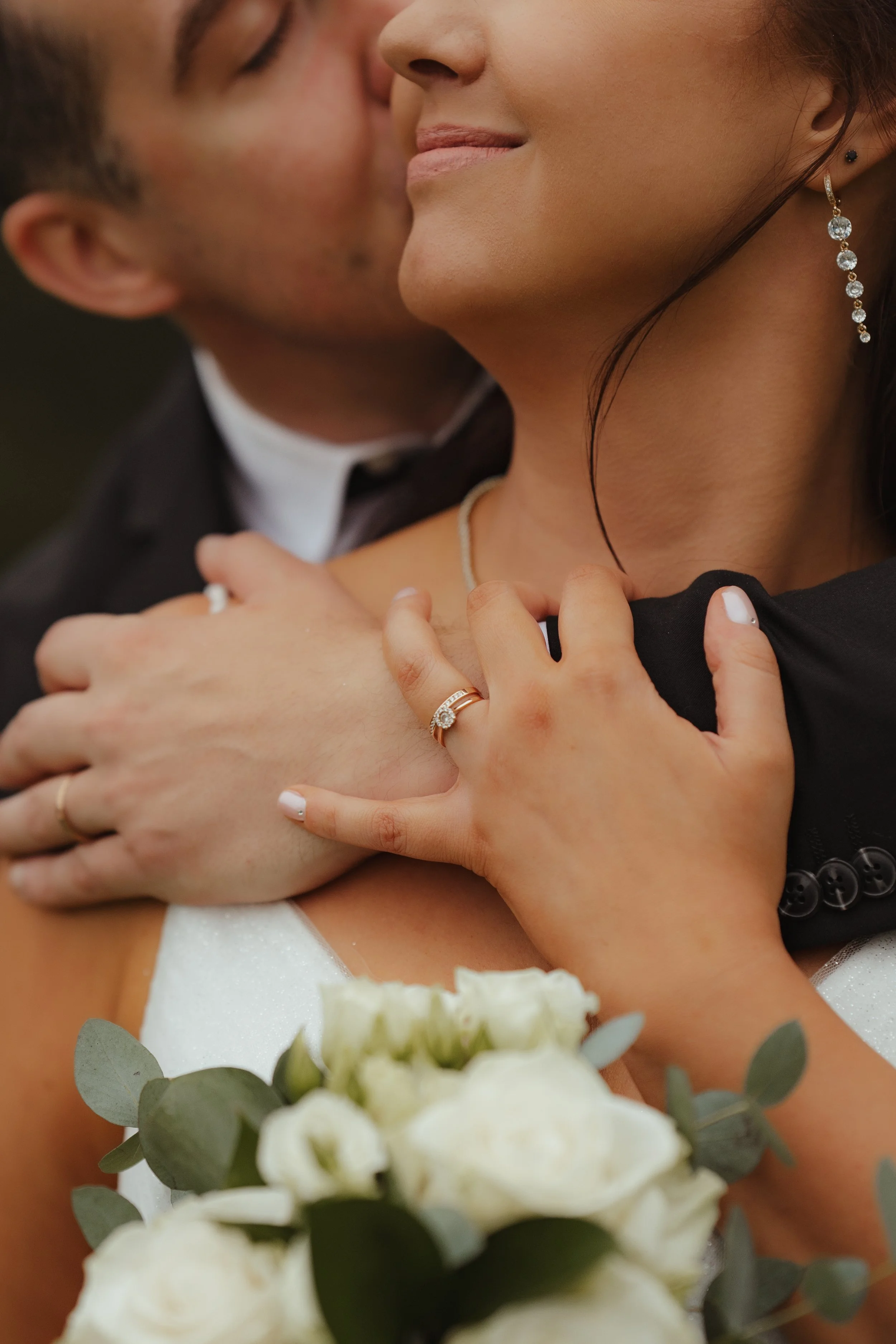 Close-up of a couple embracing, with the woman holding a bouquet of white roses. The woman is wearing jewelry, including a diamond ring and chandelier earrings, while the man is kissing her cheek.