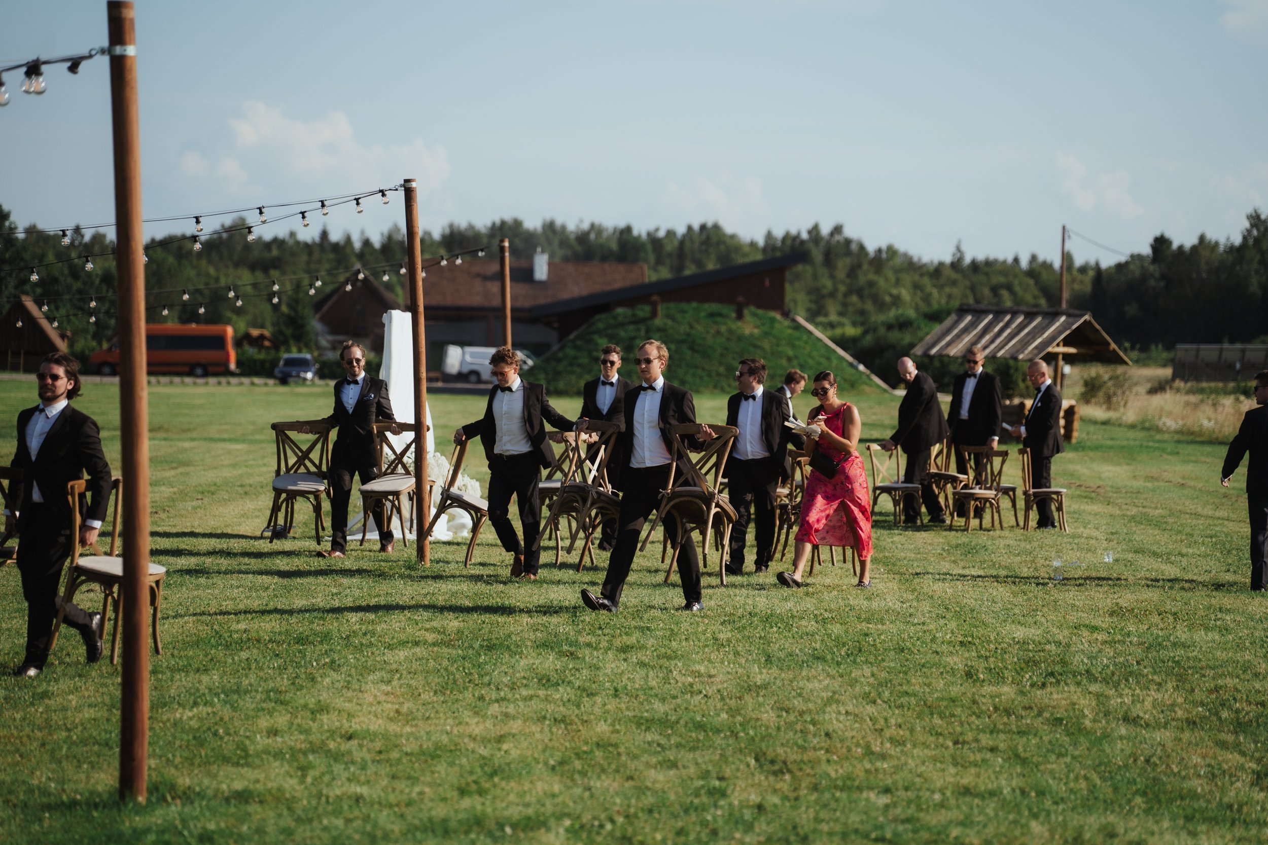 A group of men in tuxedos and a woman in a red dress setting up chairs on a grassy outdoor area for a wedding or event.