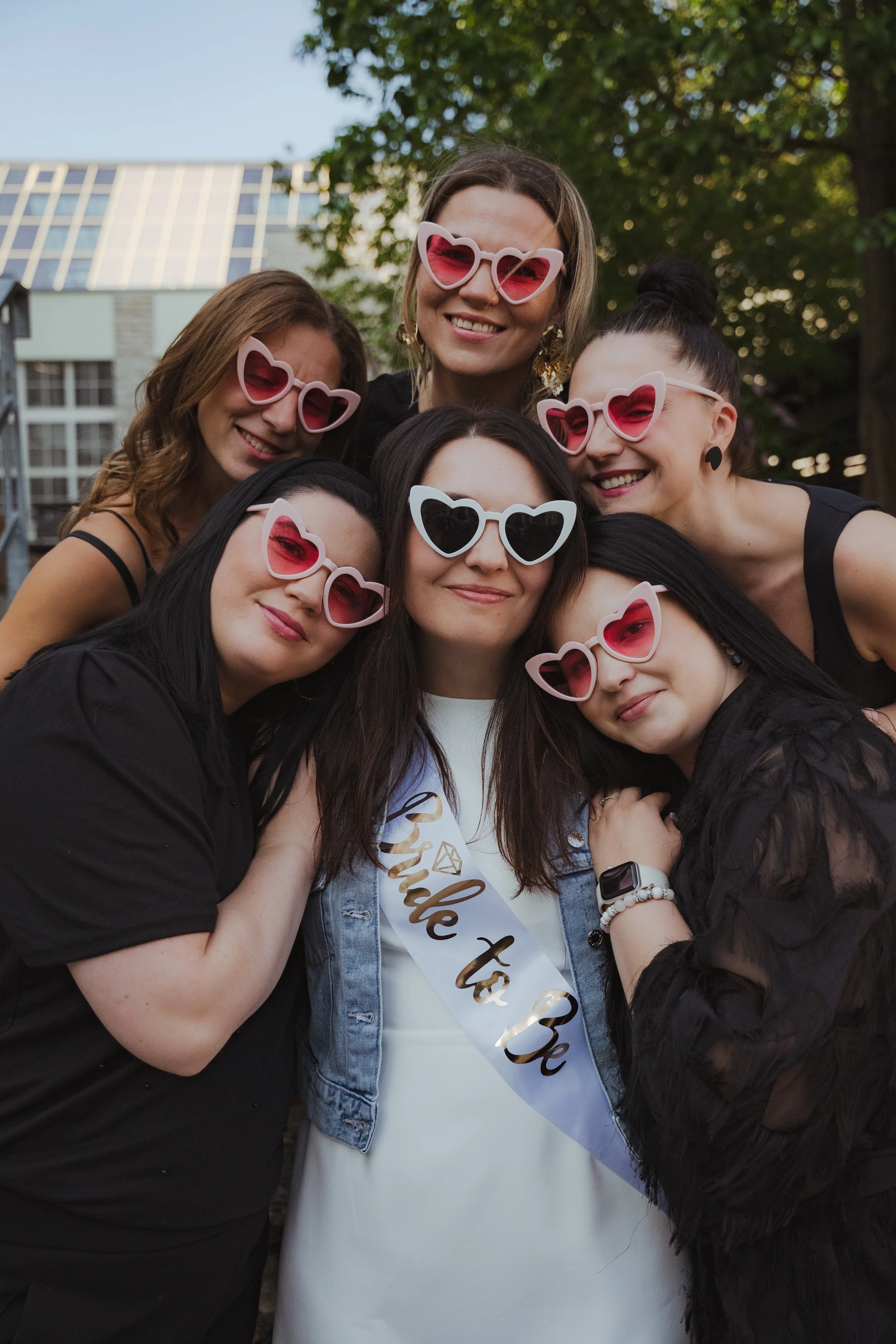 A group of women wearing heart-shaped sunglasses, celebrating outdoors with a woman in a white dress and a sash that says 'Bride to Be'.