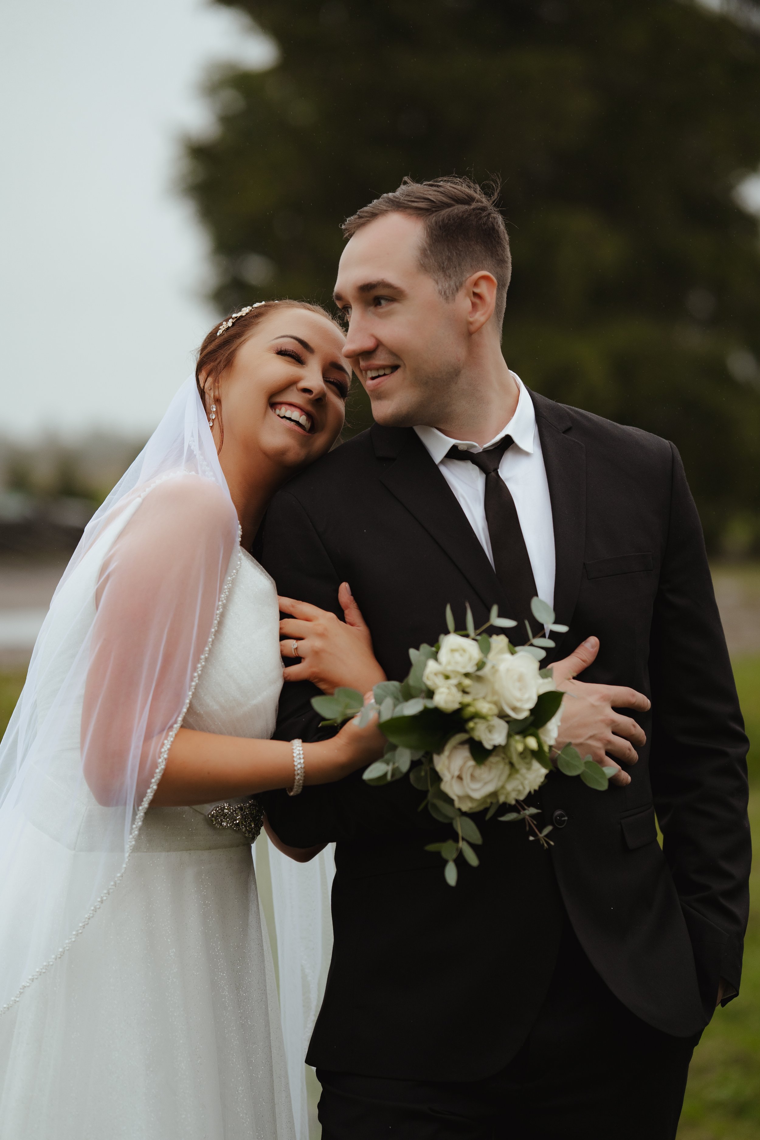 A happy bride and groom smiling at each other outdoors, the bride holding a bouquet of white roses and greenery, both dressed in wedding attire.