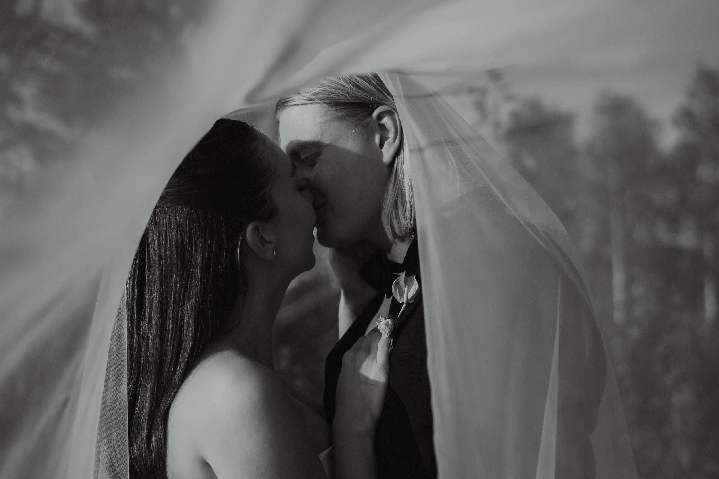 Black and white photo of two women kissing, one in a wedding dress and the other in a tuxedo, under a bridal veil outdoors.