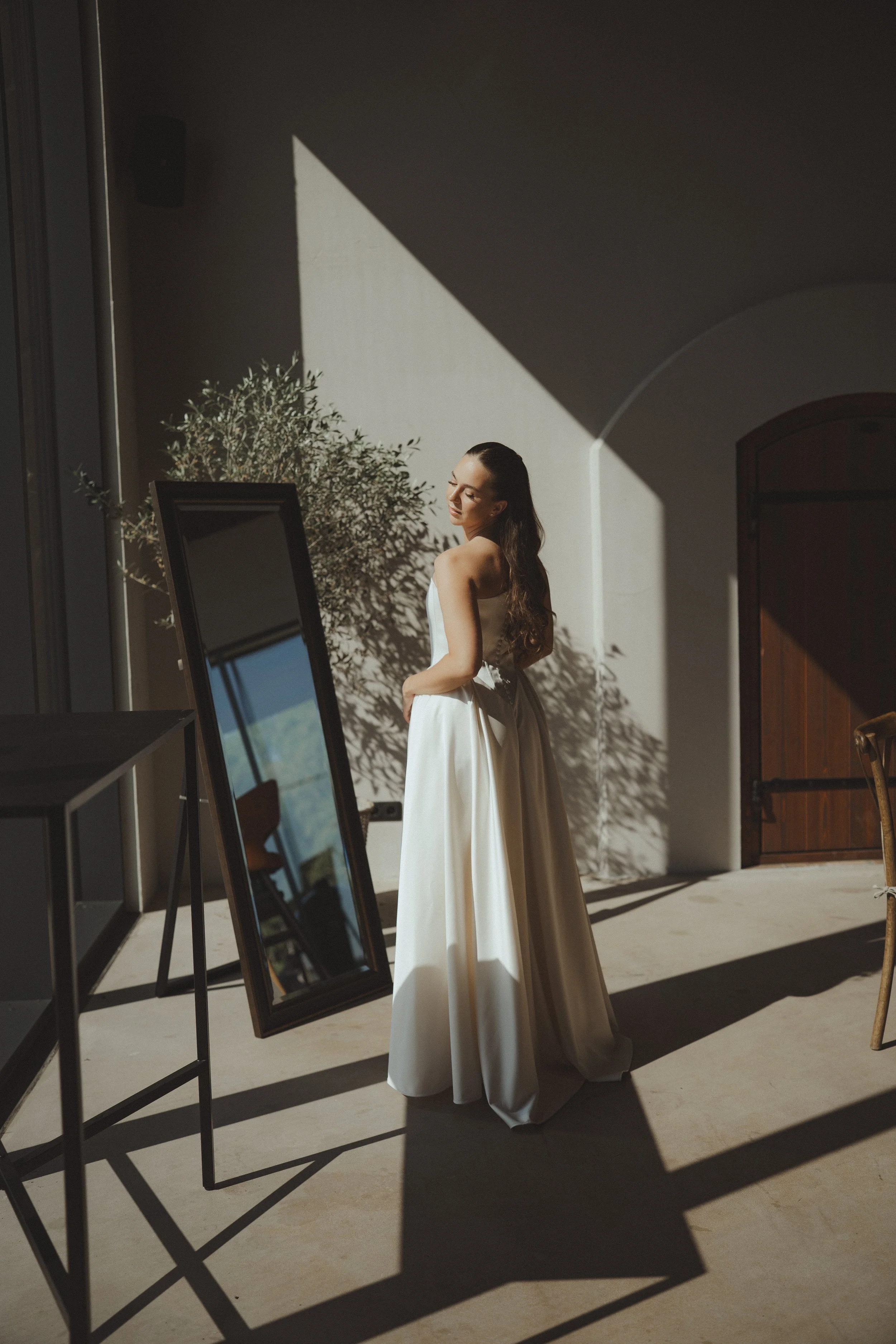 A woman in a white dress standing in a sunlit room with a large mirror, a wooden door, and a potted plant.
