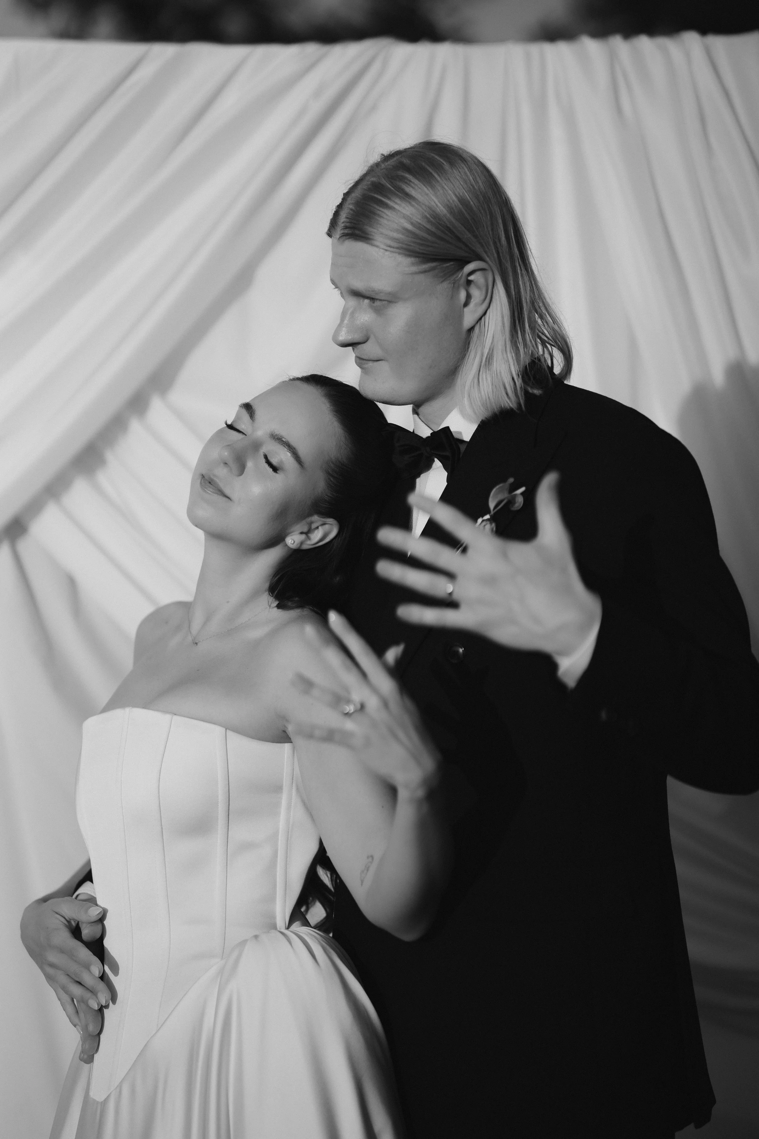 A black and white photo of a couple dancing closely; the woman is in a strapless dress with her eyes closed, and the man, with long hair, is in a tuxedo with a bow tie, holding her as they dance.