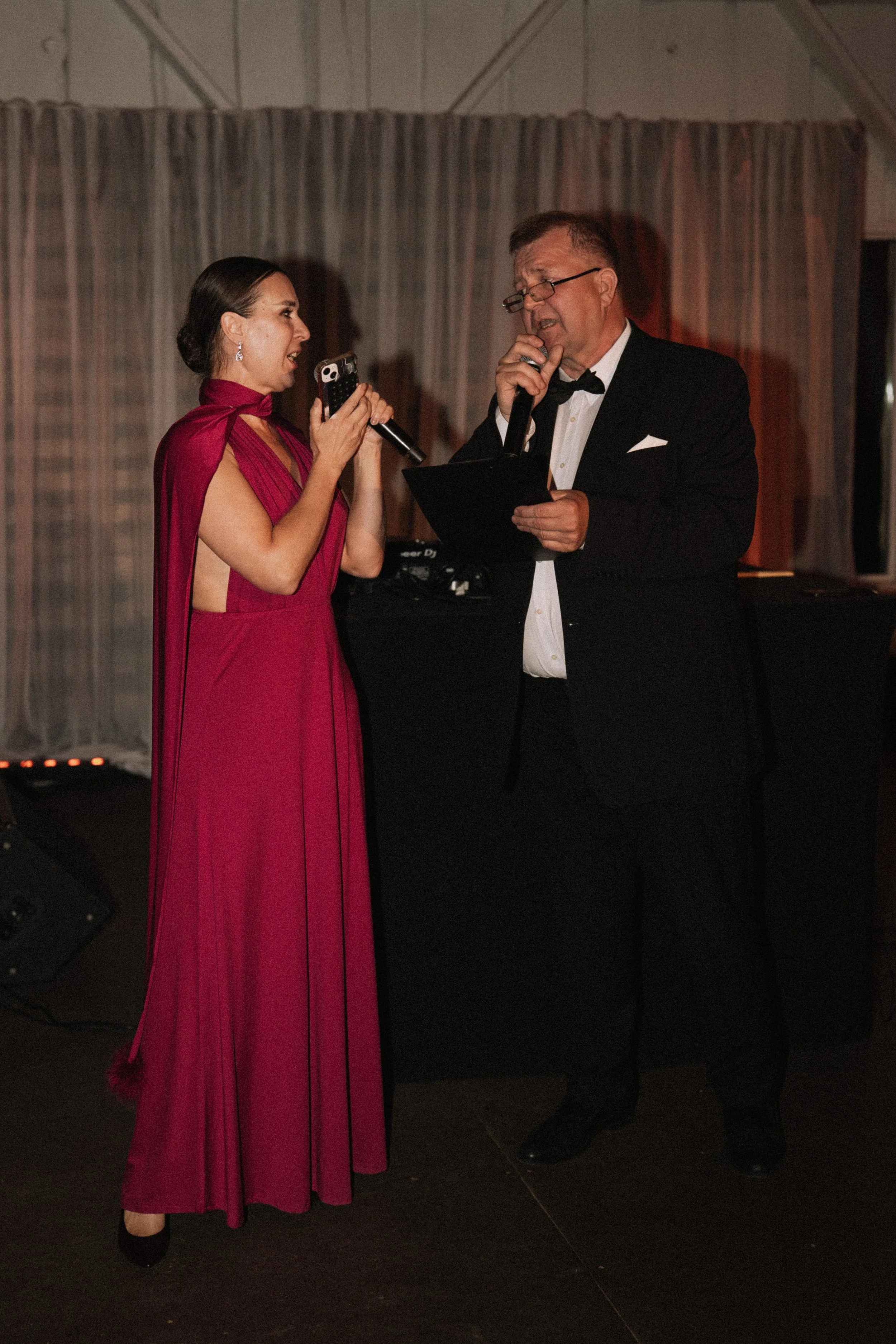 A woman in a pink gown and a man in a black tuxedo are singing into microphones at an event, with curtains in the background.