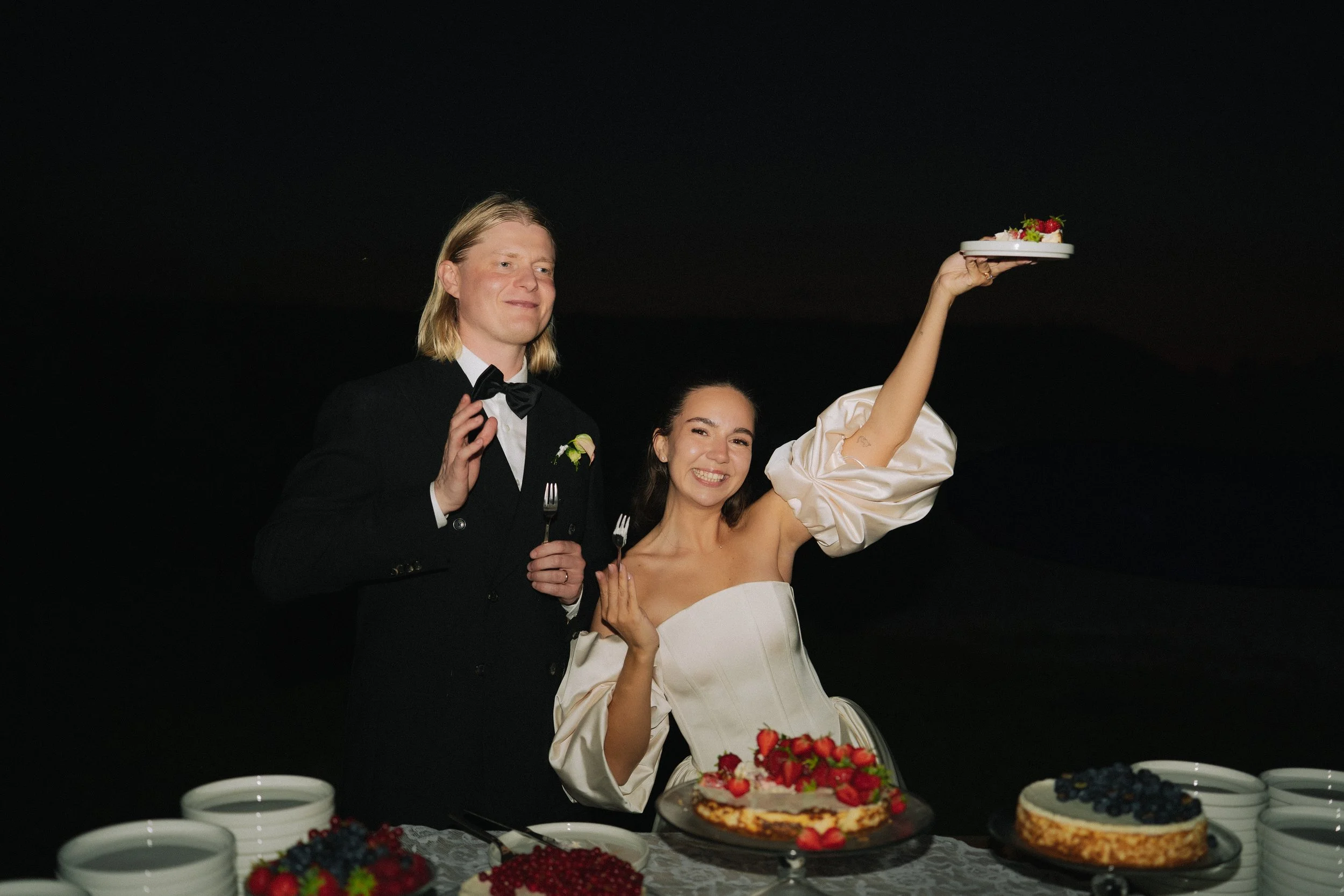 A bride and groom at a wedding celebration standing behind a table with cakes and berries, with the bride holding a plate of cake up for a toast.