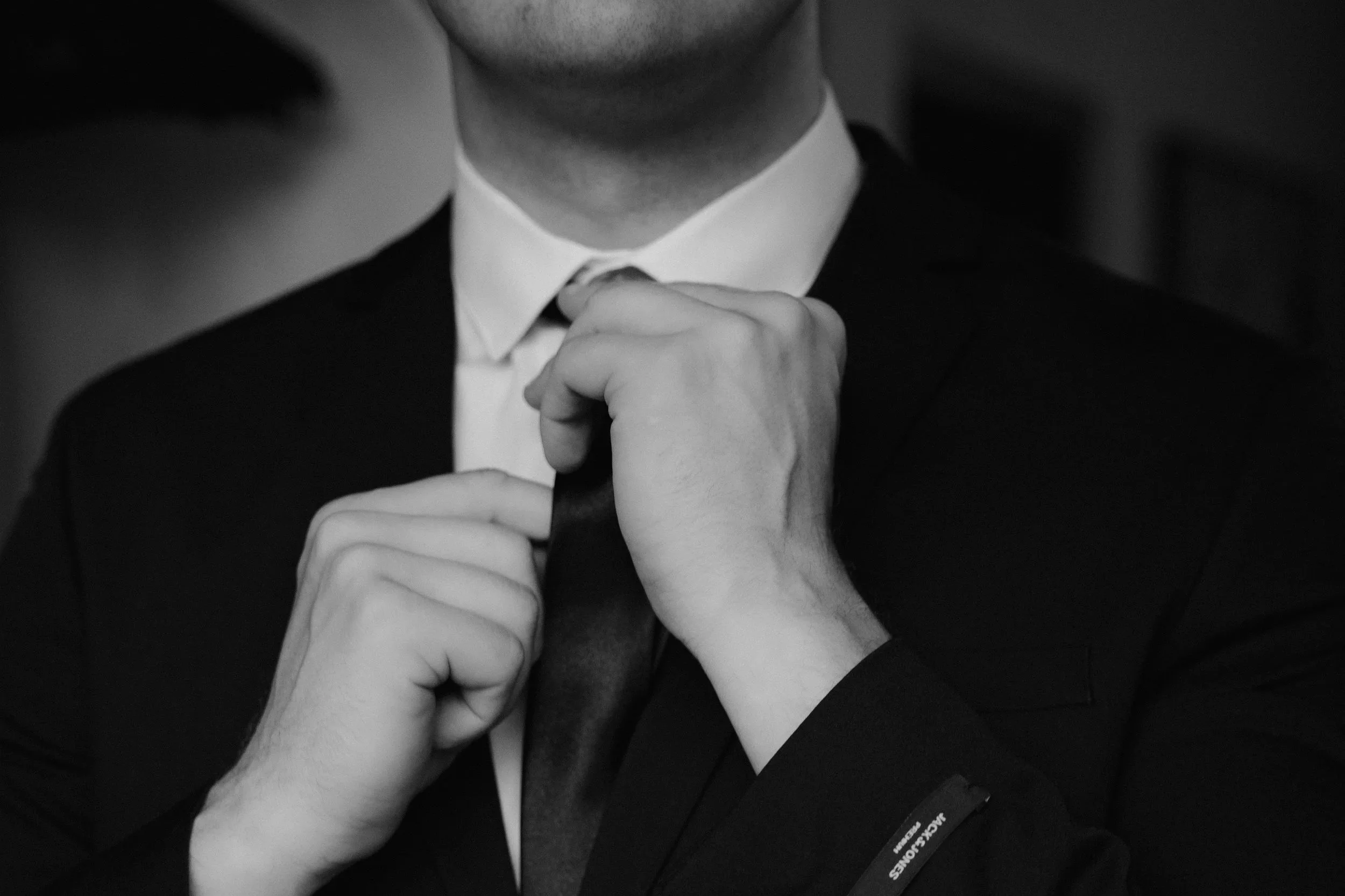 A man adjusting his tie while wearing a suit and white dress shirt in black and white.