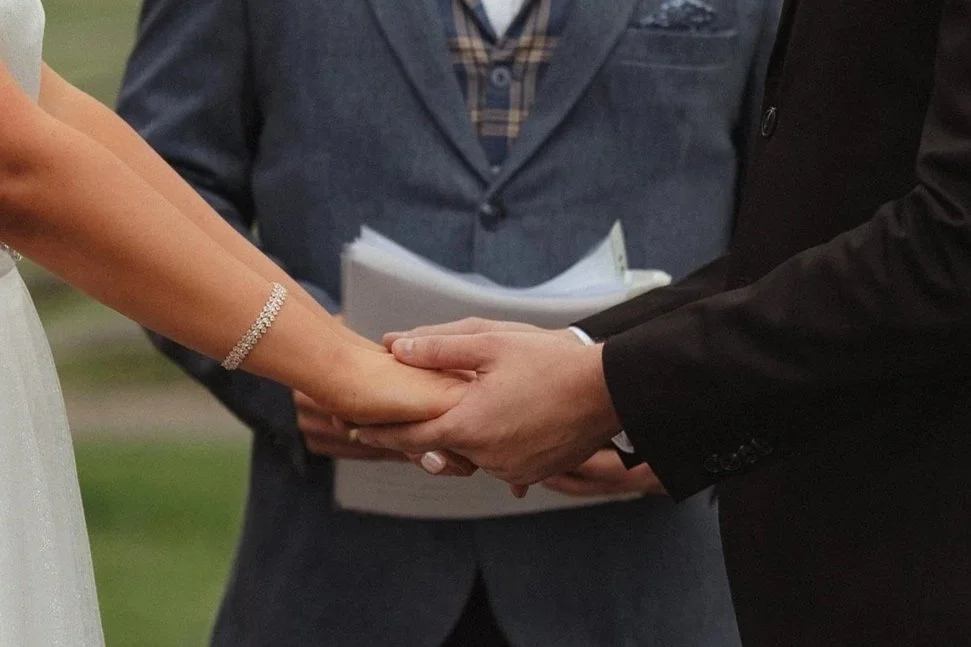 A close-up of a couple holding hands during a wedding ceremony, with an officiant standing behind them holding a book and papers.