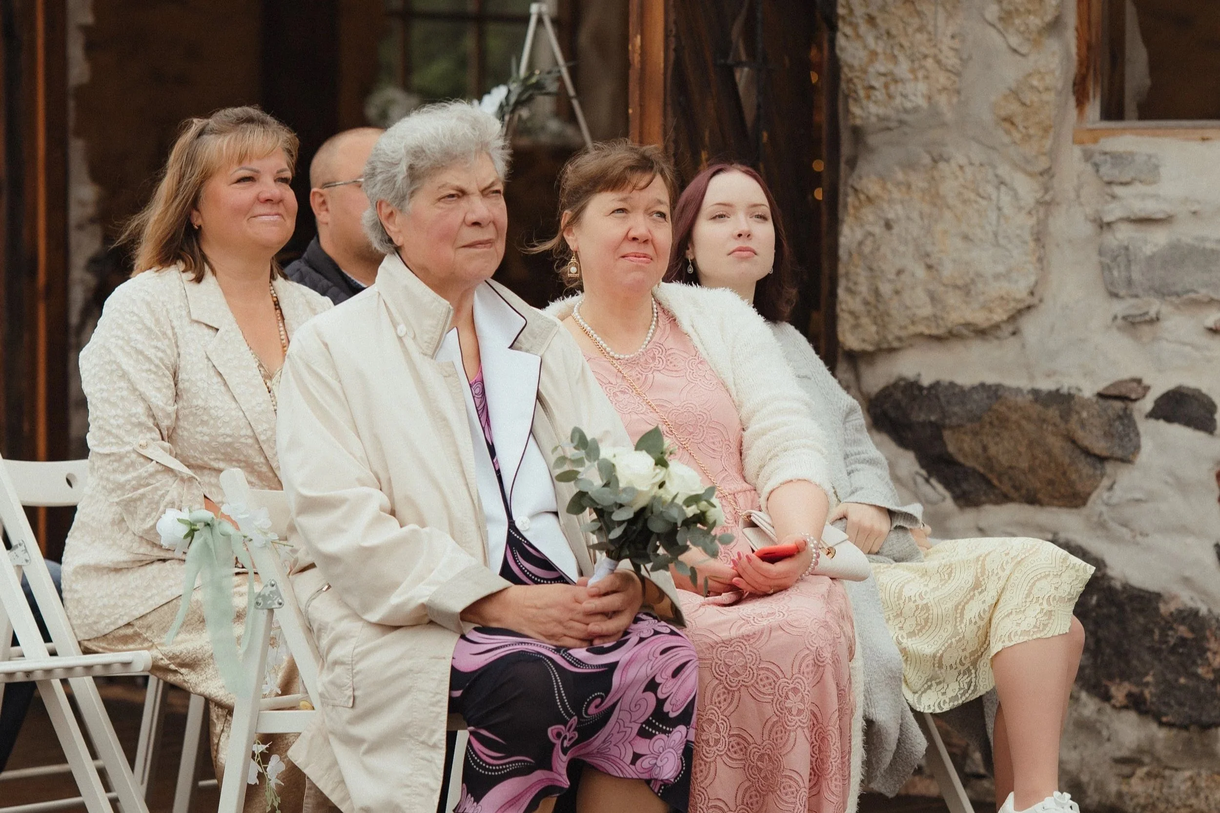 Group of women sitting at a gathering or ceremony, seated in chairs outdoors near a stone and wood structure, with one woman holding a small bouquet of white flowers.