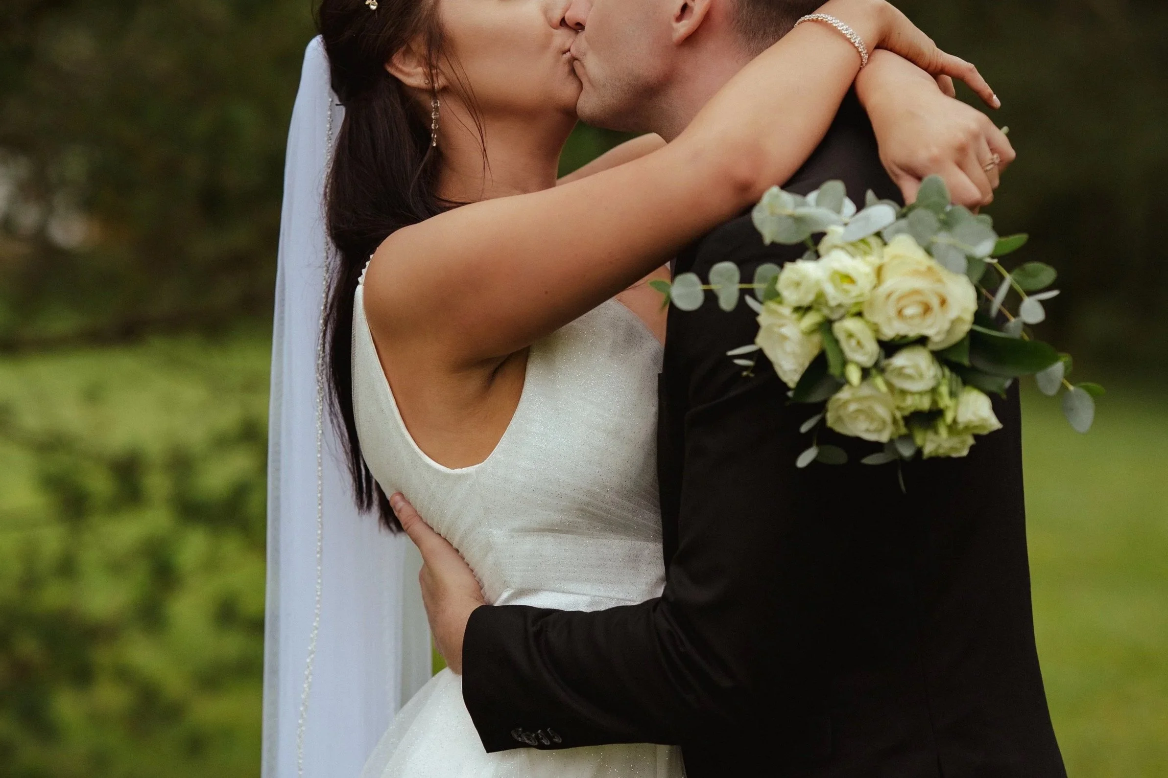 A bride and groom kissing outdoors, with the bride holding a bouquet of white roses and greenery.