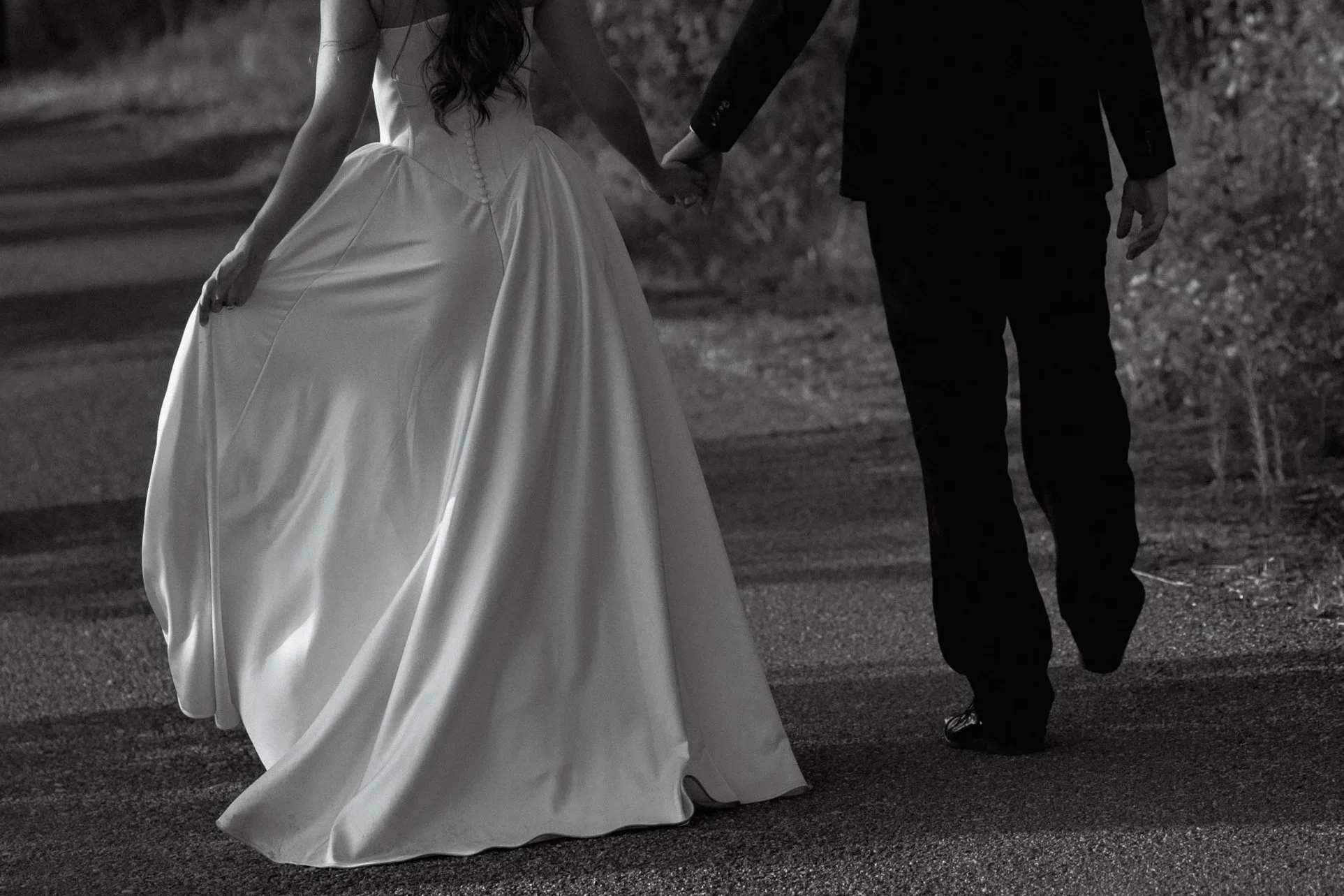 Black and white photo of a couple walking hand-in-hand on a road, the woman in a wedding dress and the man in a tuxedo.