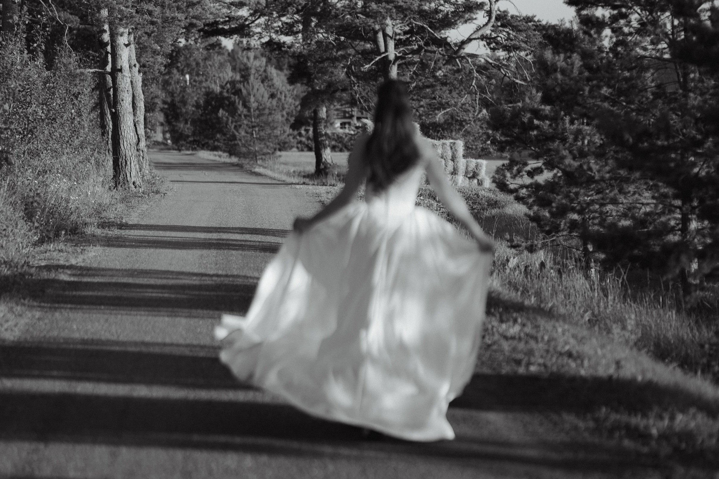 A woman in a long, flowing white dress walking along a rural gravel road lined with trees and hay bales, with her back turned to the camera.