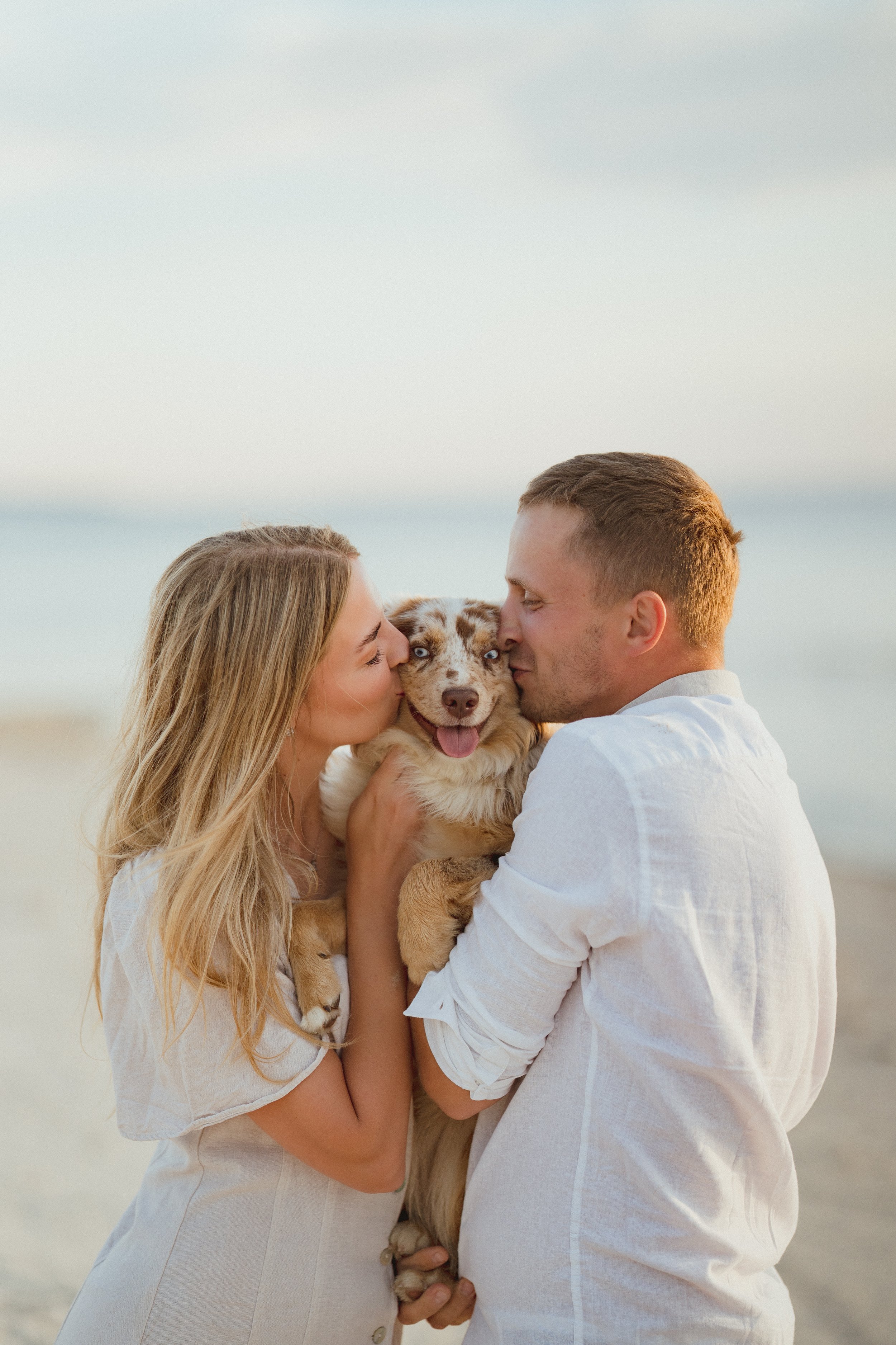 A couple kisses their dog on the beach during sunset.