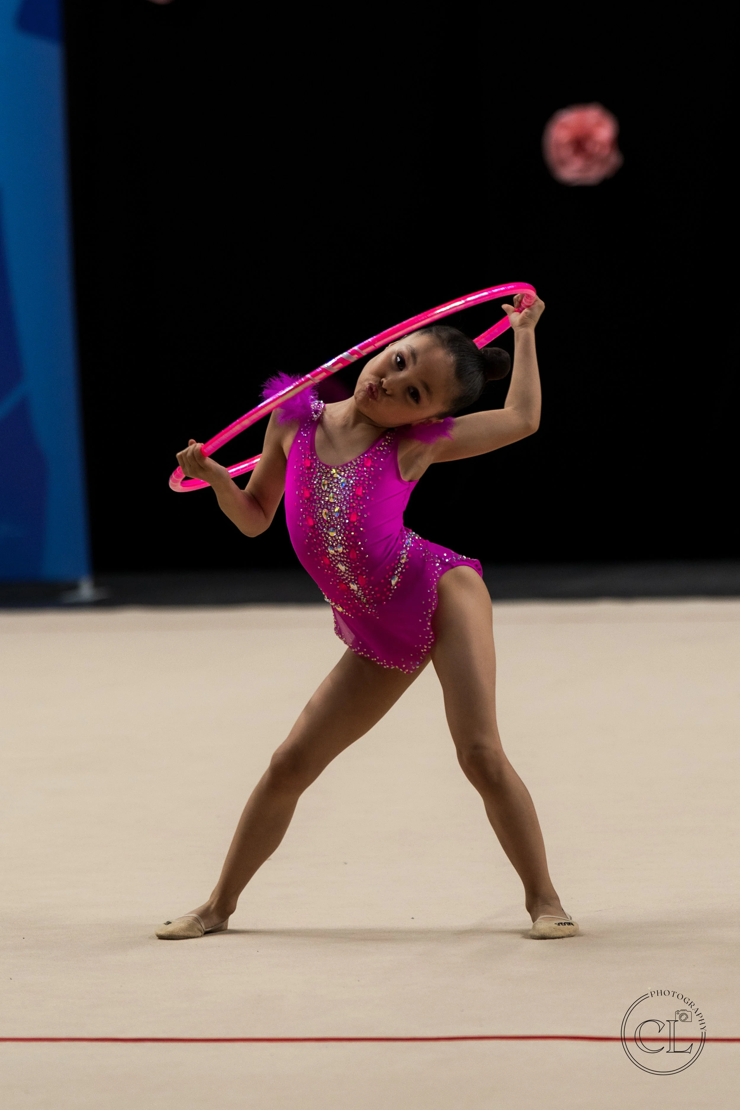 Young gymnast in a pink leotard with rhinestone embellishments practicing rhythmic gymnastics with a pink hoop on a beige gym floor.