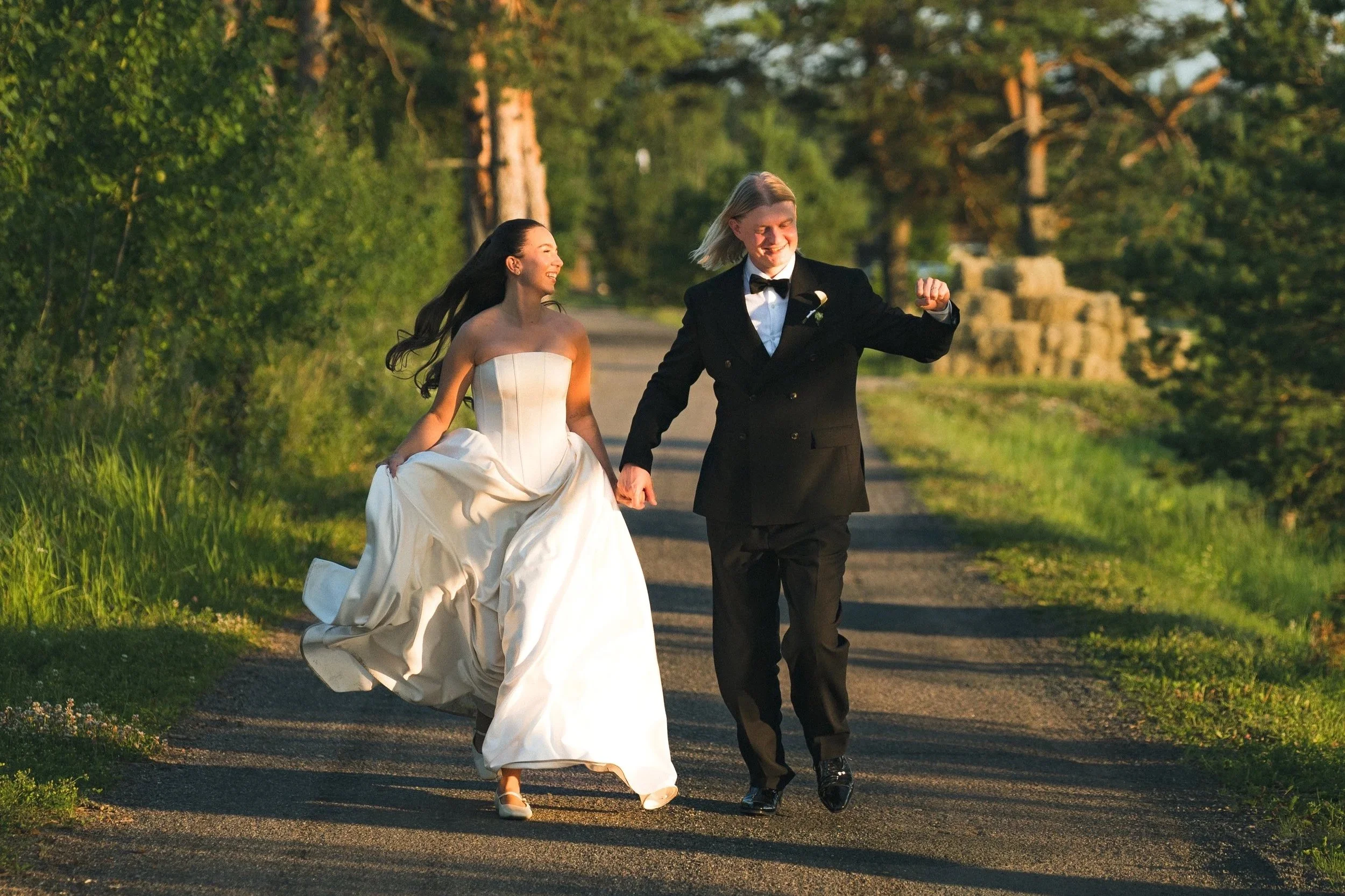 A newlywed couple walking and holding hands on a country road during sunset. The bride is in a strapless white wedding dress with a full skirt, and the groom is in a black tuxedo with a bow tie.