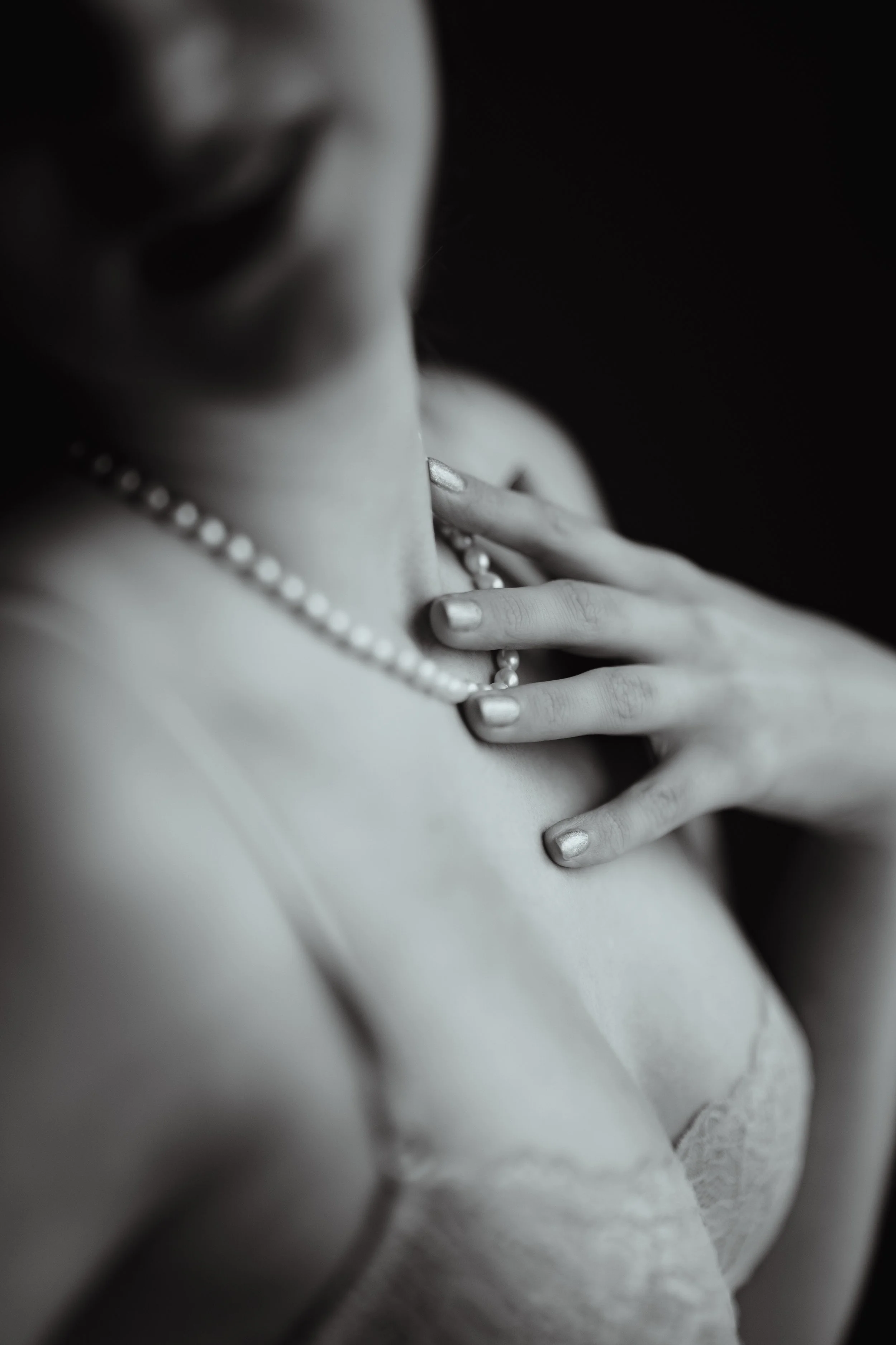 Close-up black and white photo of a woman's hand resting on her chest, wearing a pearl necklace, with her face partially out of frame.