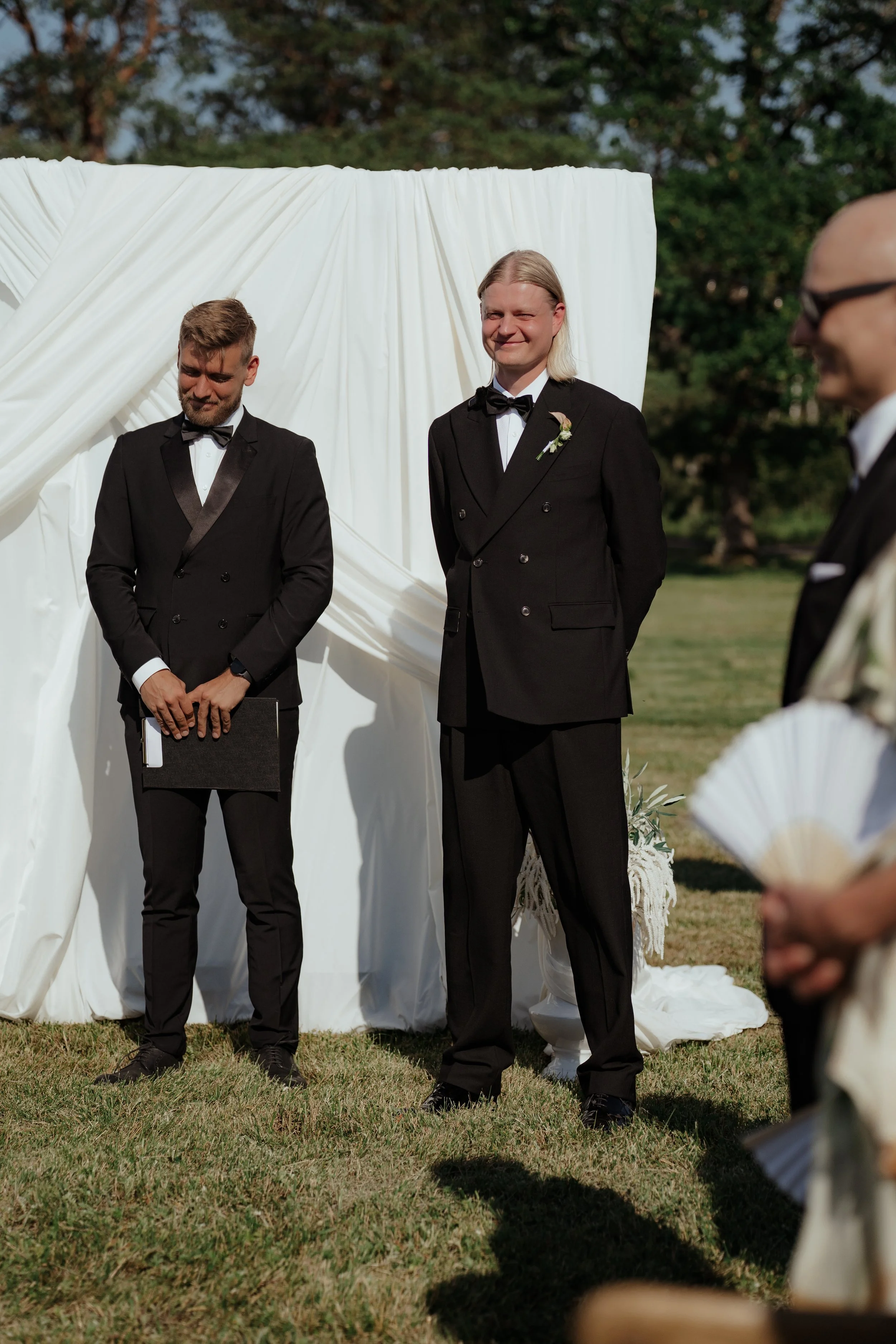 Two men in tuxedos standing outdoors during a wedding ceremony, one with long blonde hair smiling and the other with short hair looking down, with a white fabric backdrop behind them.