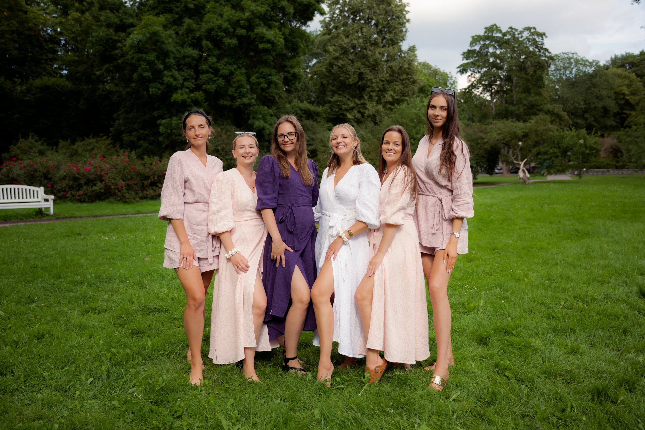 Six women standing in a park on the grass, dressed in colors and styles suitable for a summer day, smiling for the camera with trees and a park bench in the background.