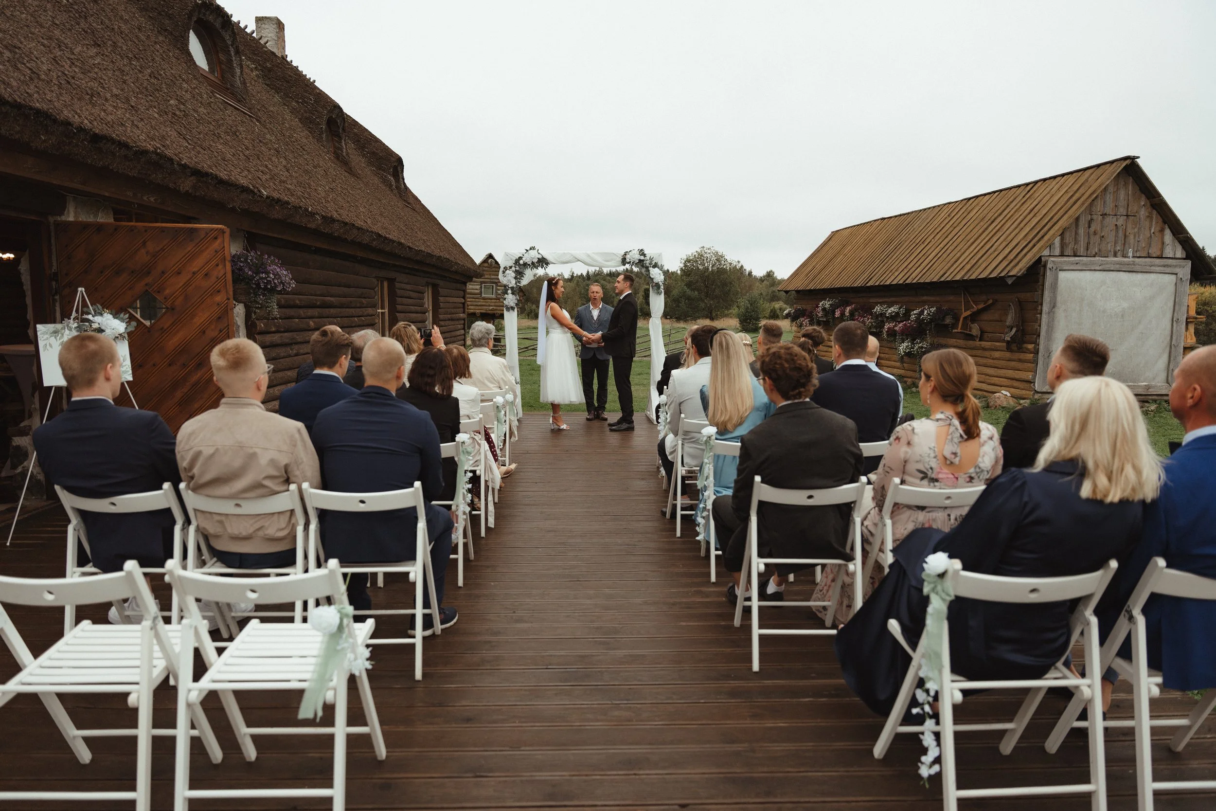An outdoor wedding ceremony with a bride and groom exchanging vows under a white floral arch, surrounded by seated guests on a wooden deck, with rustic buildings and green trees in the background on a cloudy day.