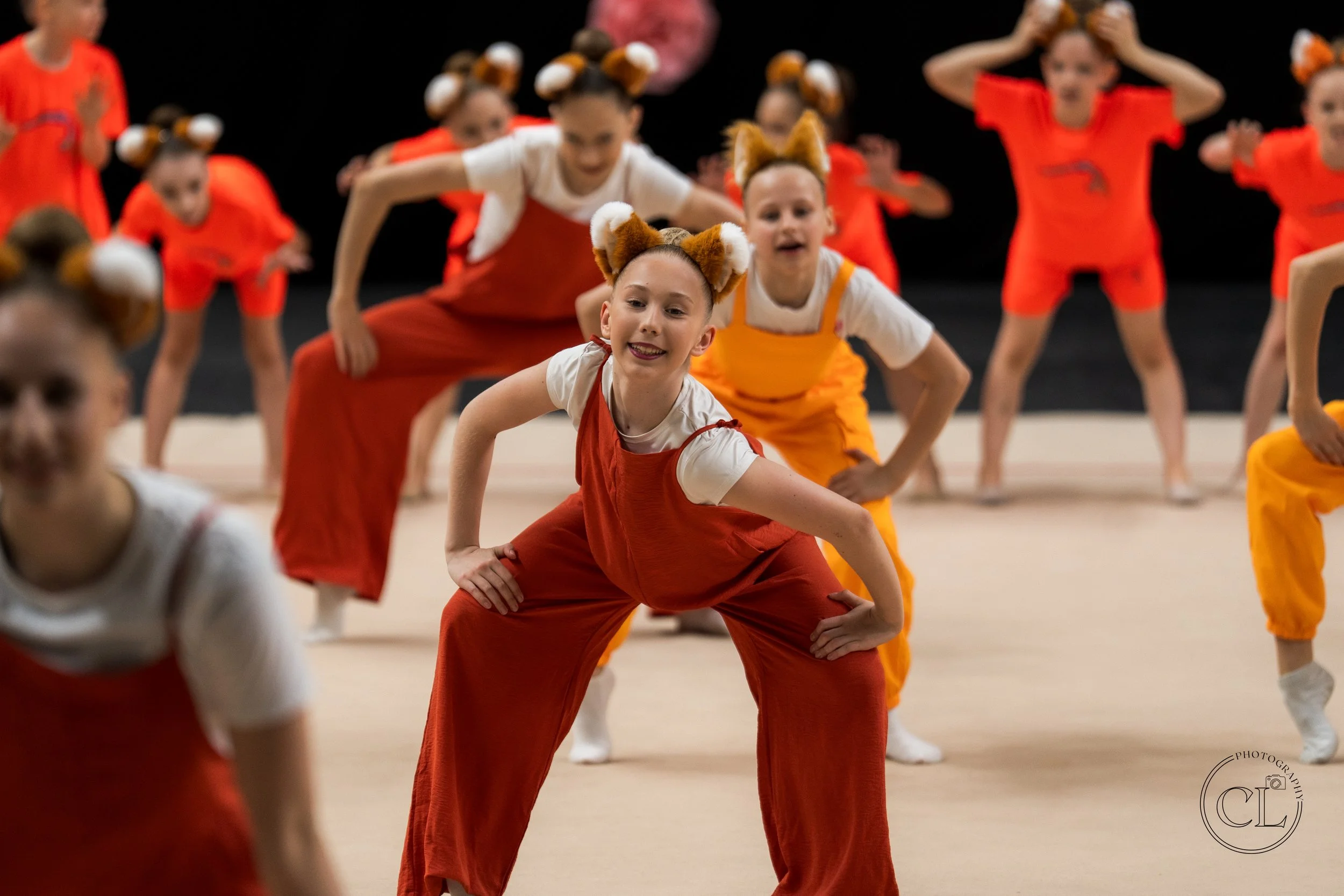 Group of young girls dressed in colorful costumes with animal ear headbands, performing a dance routine on stage.