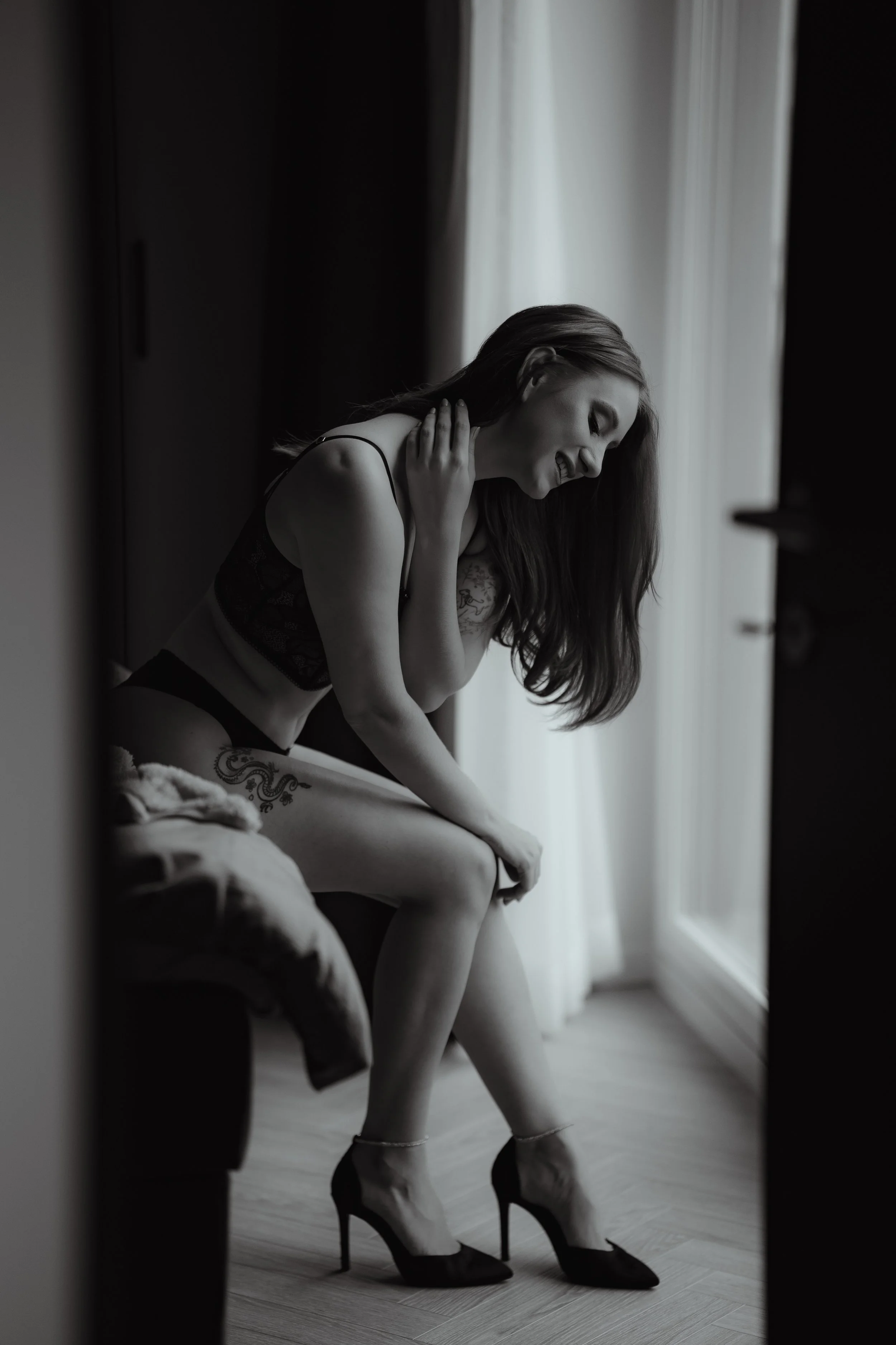 A woman in lingerie and high heels sitting on a bed near a window, smiling and touching her neck, in black and white.
