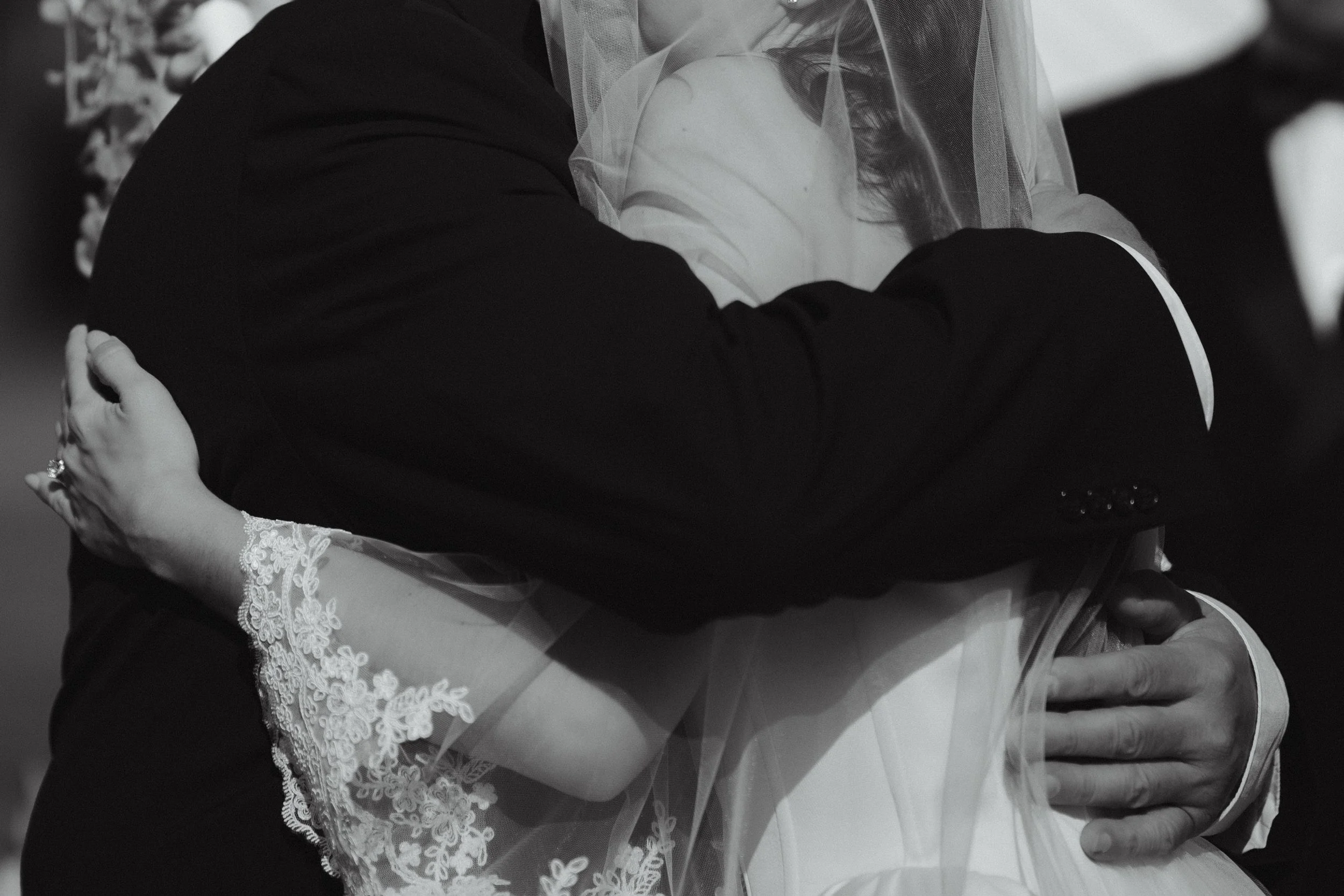A couple hugging tightly on their wedding day, with the bride in a dress with lace details and a veil, and the groom in a dark suit.