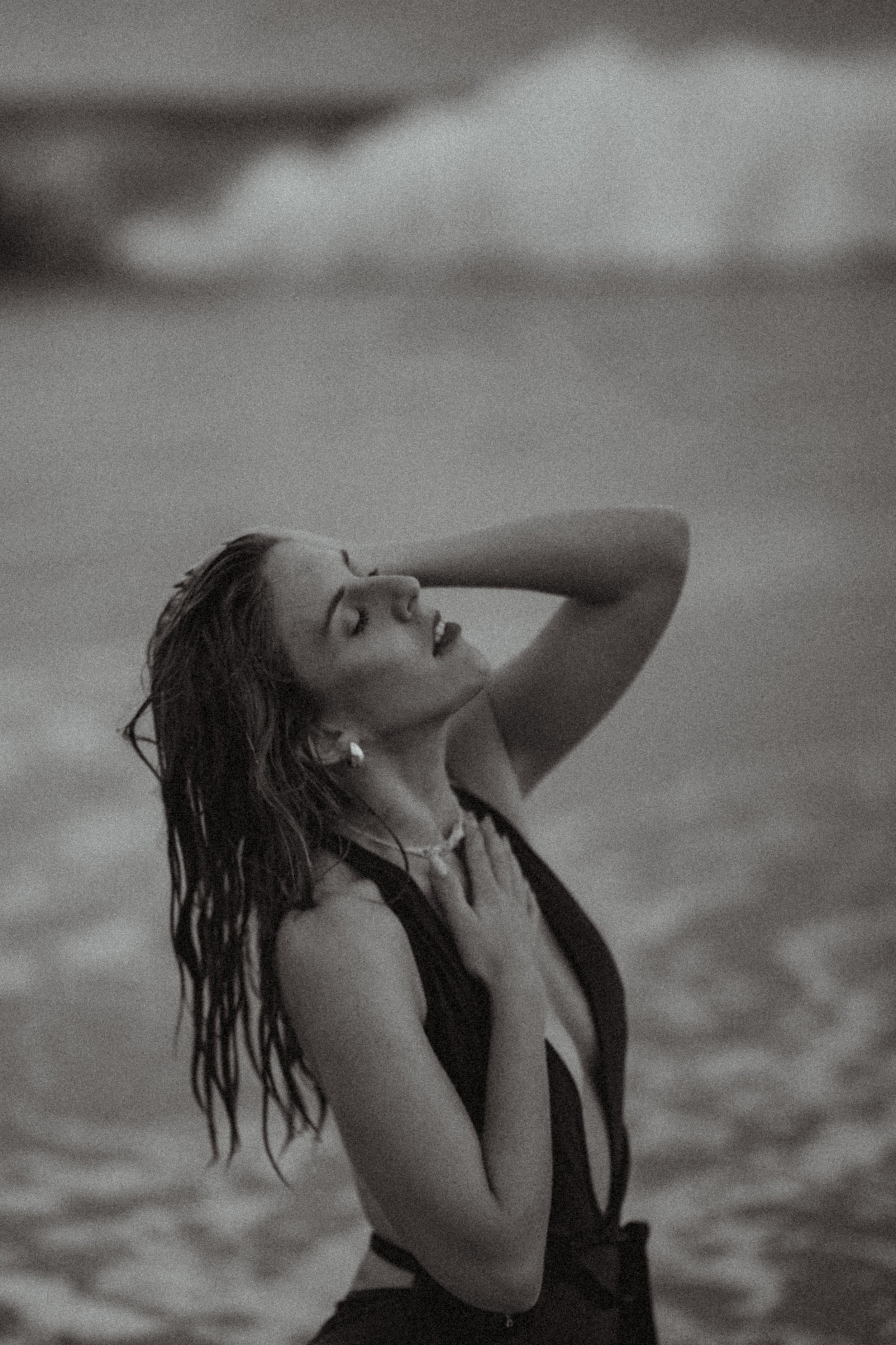 A black and white photo of a woman with wet hair, eyes closed, head tilted back, one hand on her chest, the other resting on her head, standing in water with cloudy sky in the background.