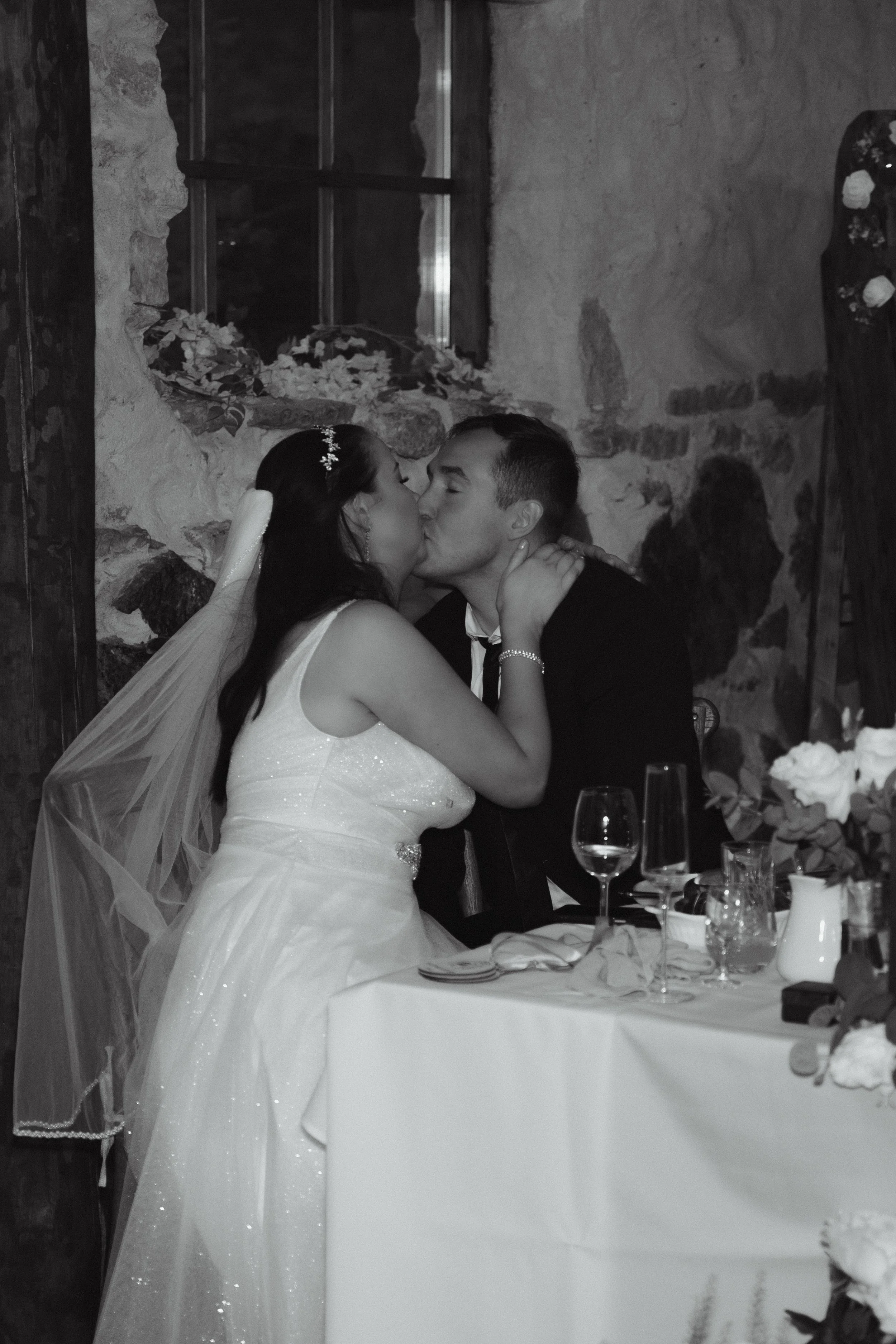 A bride and groom kissing at their wedding reception in a rustic setting with stone walls and a window.