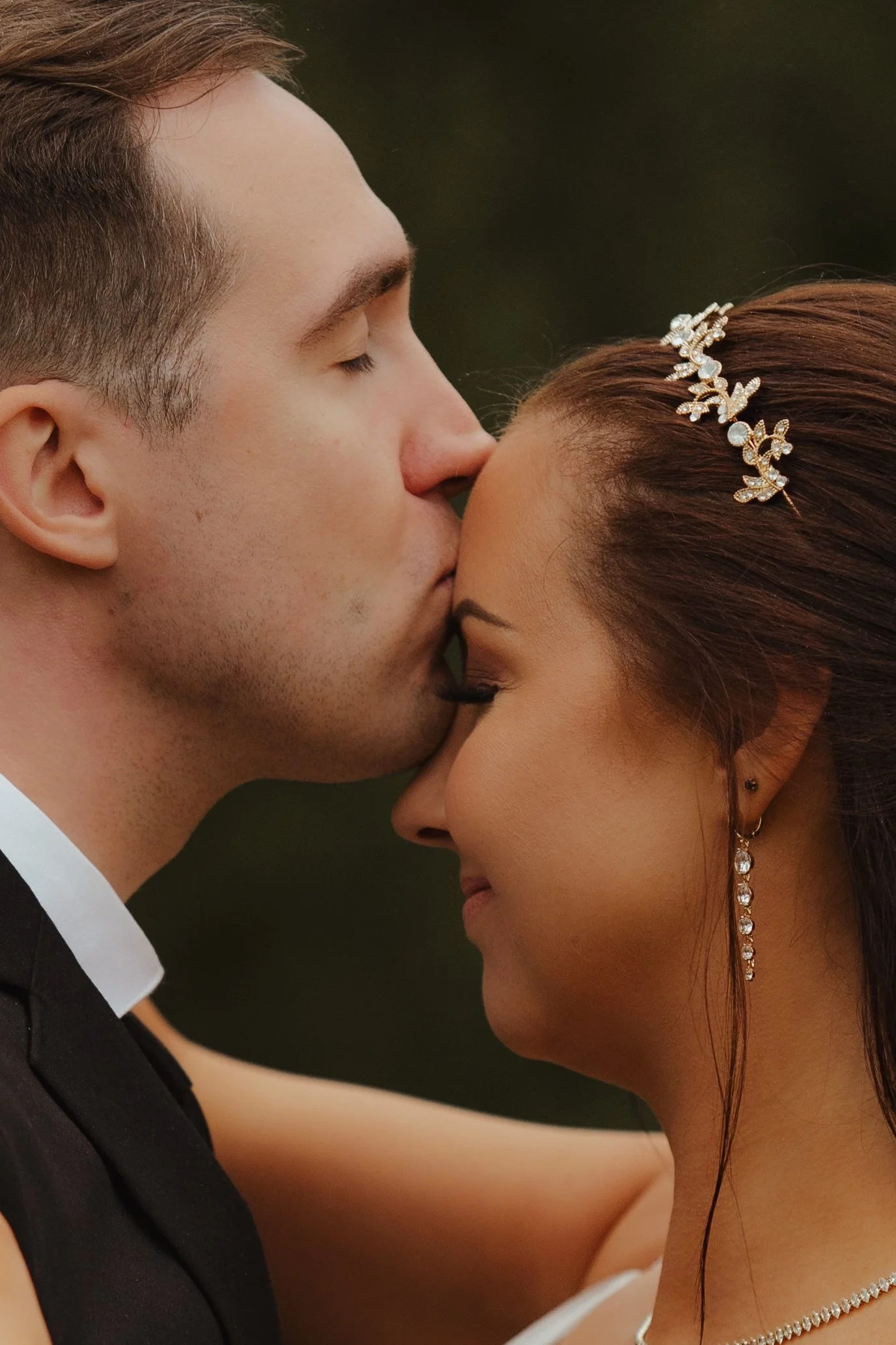 A close-up of a bride and groom with their foreheads touching, sharing an intimate moment during their wedding, with the groom kissing the bride's forehead, both with closed eyes and wearing elegant jewelry.