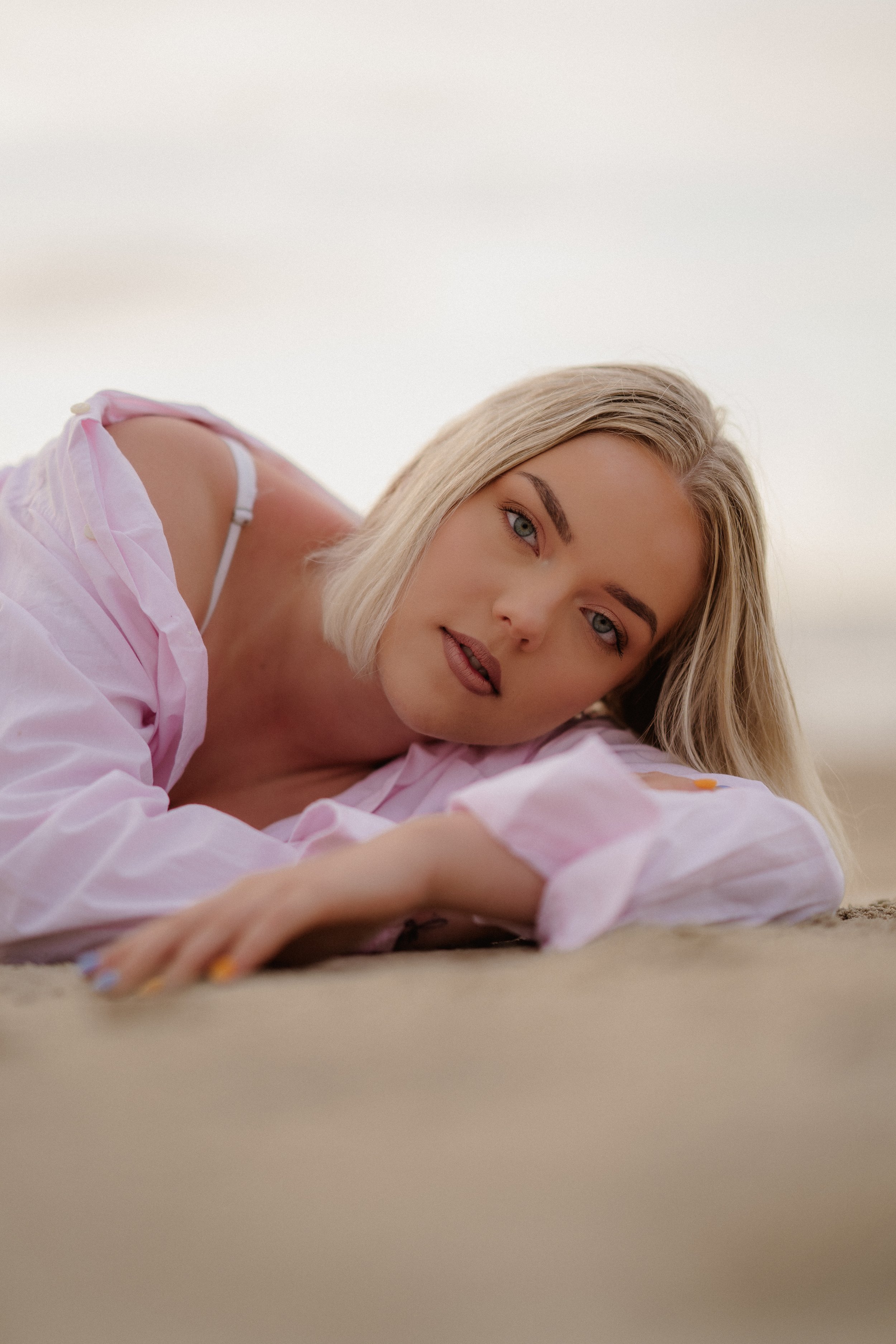 A young woman with blonde hair and blue eyes lying on the ground, wearing a pink shirt and a white bra strap visible.