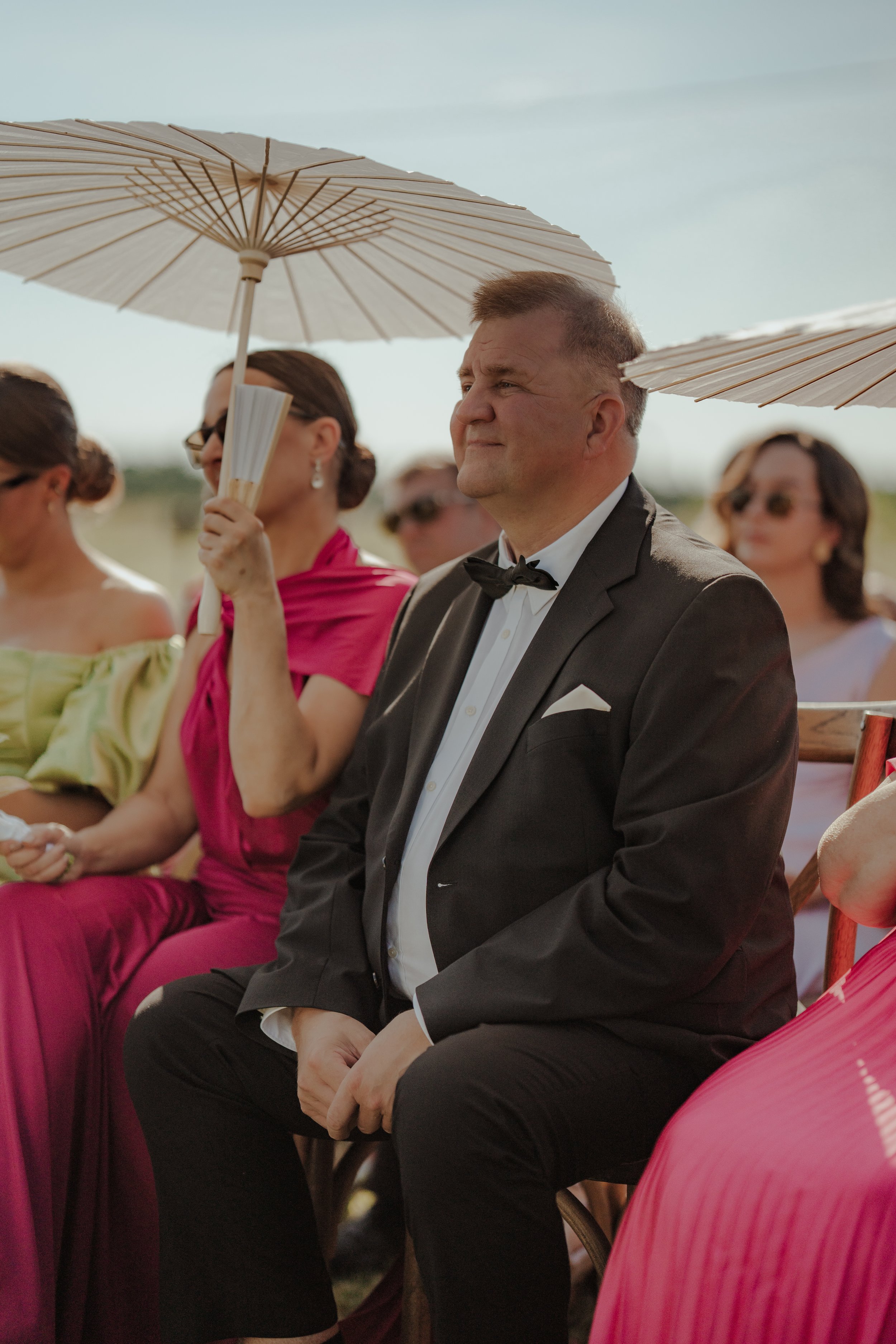 A man in a black tuxedo with a bow tie, sitting outdoors at an event, holding a paper fan, with women using umbrellas behind him.