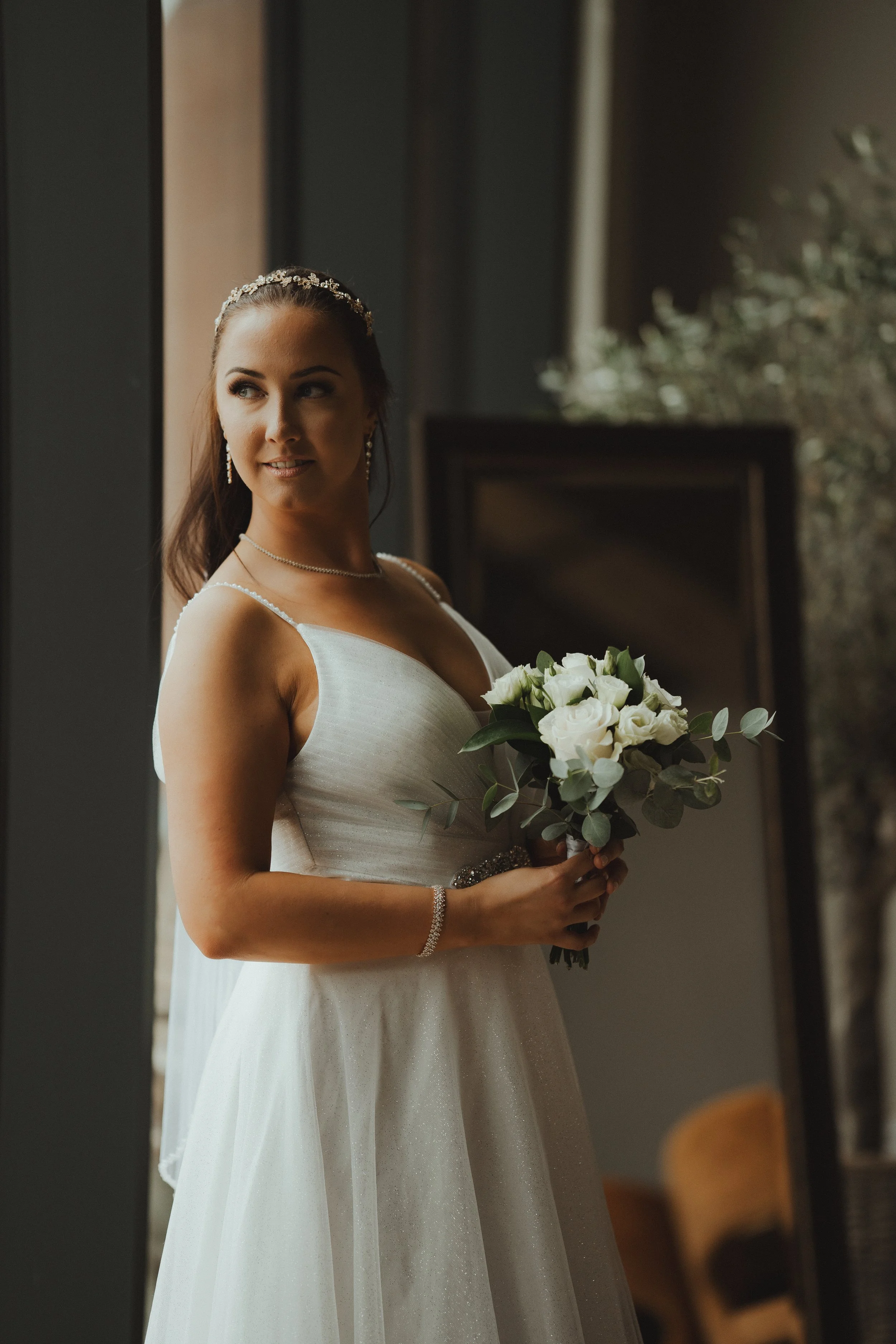 A bride in a white wedding dress holding a bouquet of white roses with green leaves, standing indoors near a window.