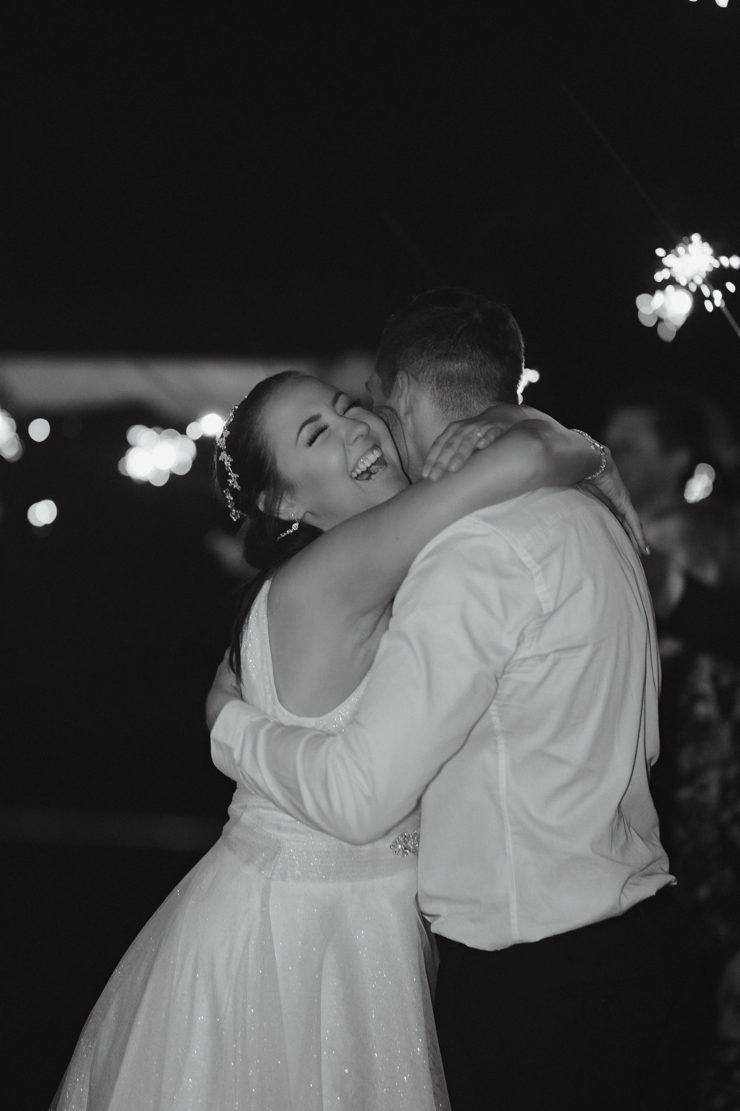 A bride and groom embrace and laugh at their wedding reception during nighttime, with blurred lights and sparklers in the background.