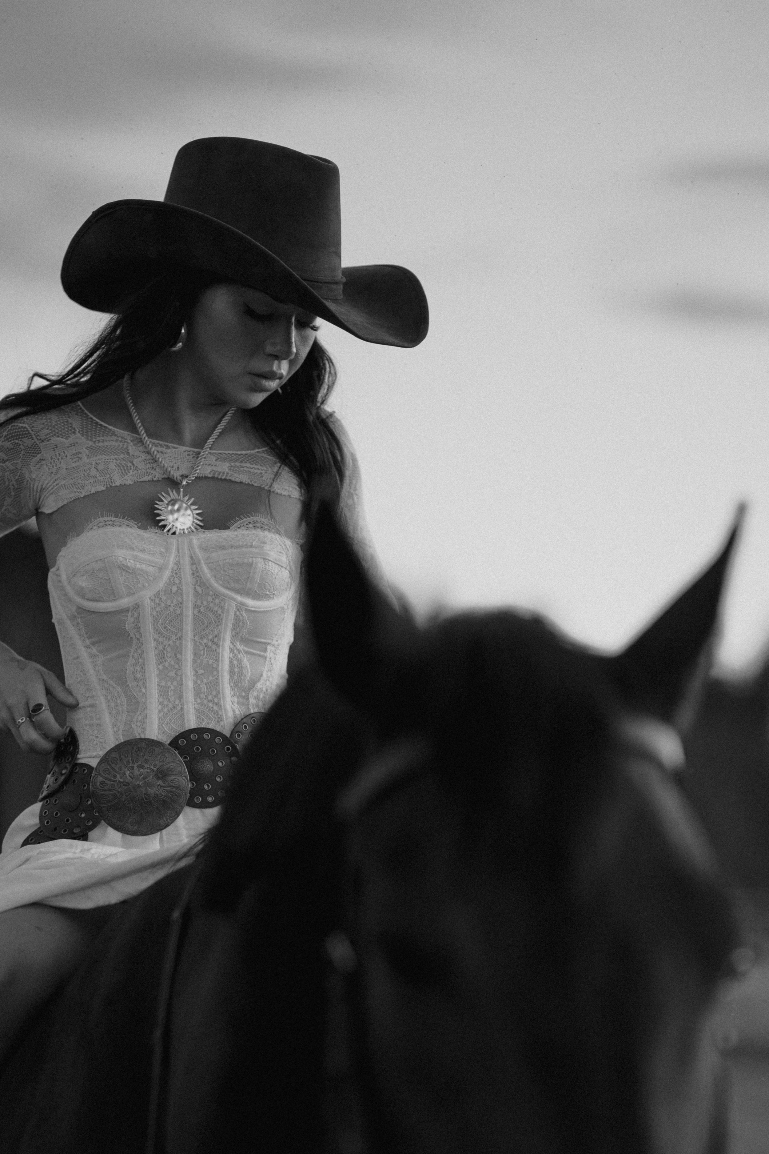 A woman wearing a large cowboy hat, lace top, and decorative belt sitting on a horse with a blurred landscape in the background.