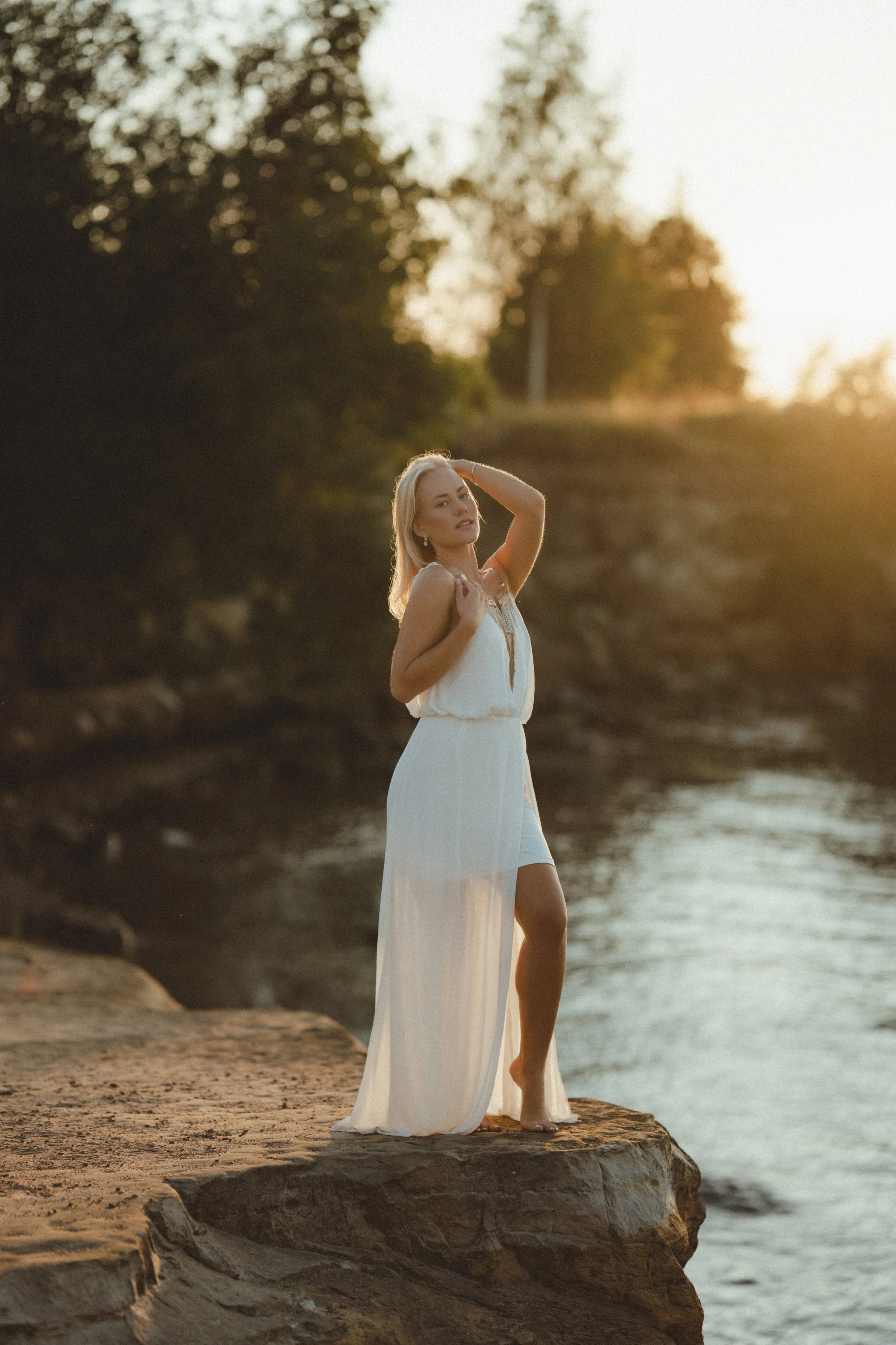 A woman in a white dress standing barefoot on a rock by a river at sunset, with trees and a bright sky in the background.
